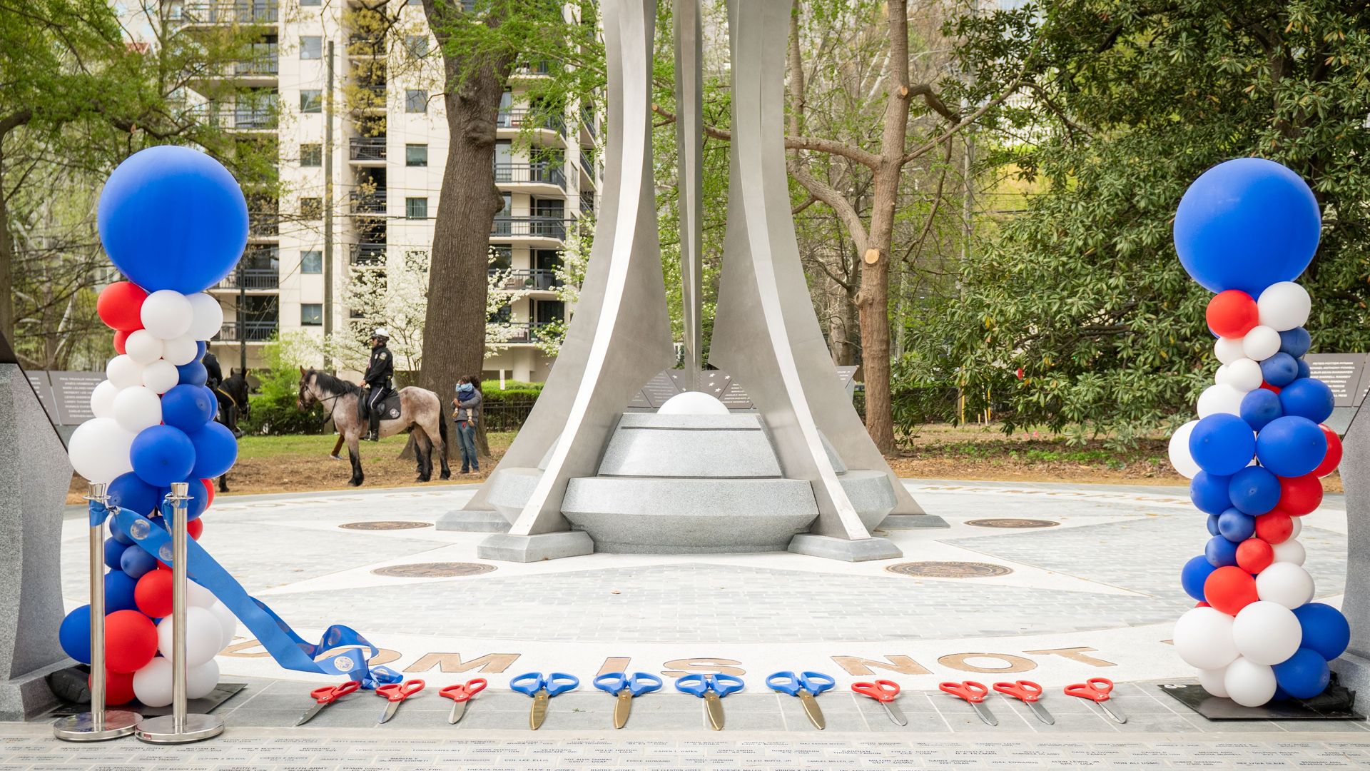 Outdoor monument with four tall curved silver pillars. Front features blue scissors and a blue ribbon on pavement; red, white, and blue balloon columns flank the scene. A horse and rider appear in the background.