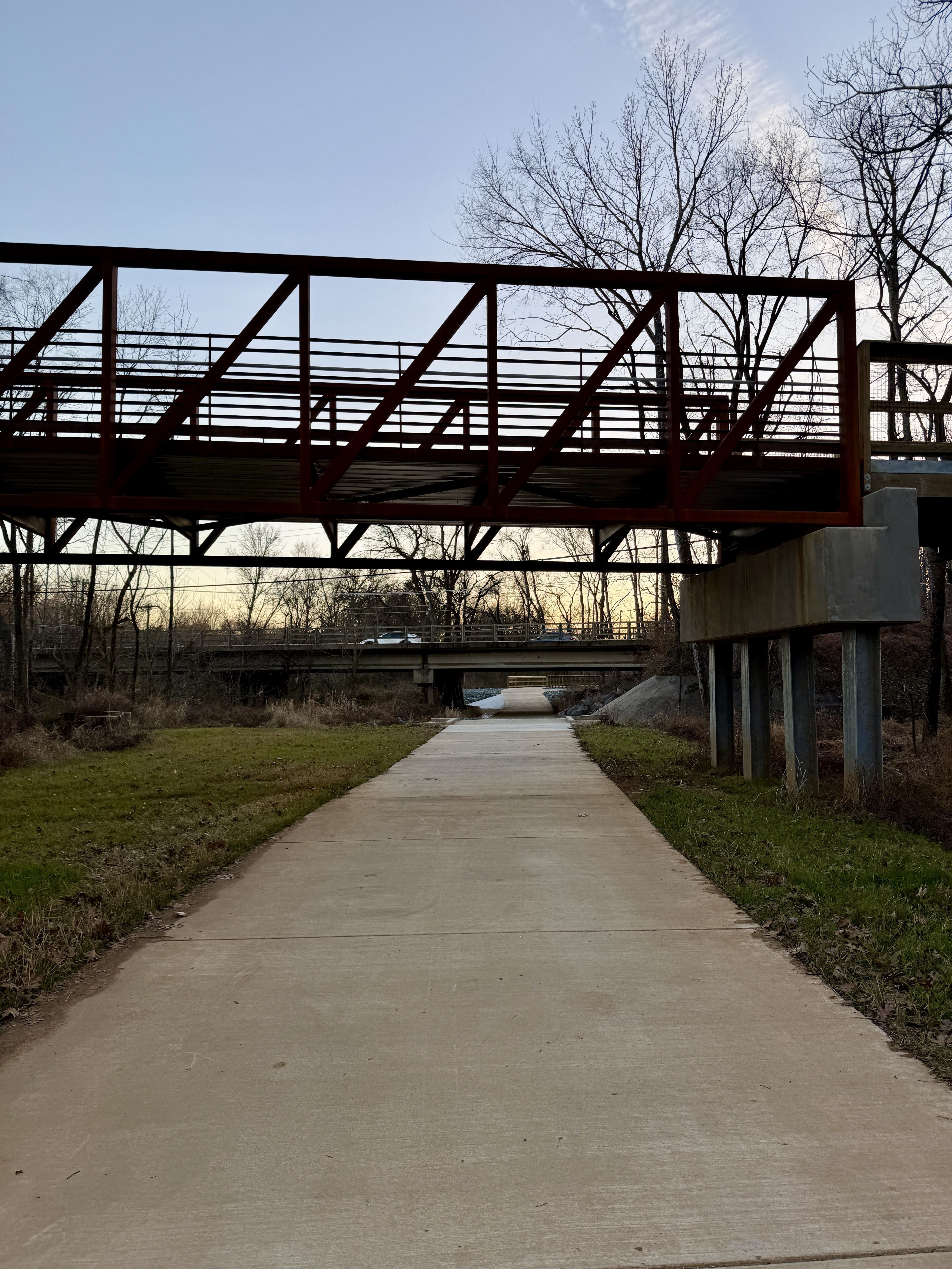Concrete path leading under a rust-colored metal pedestrian bridge with bare trees around, and a sunset sky in the background.