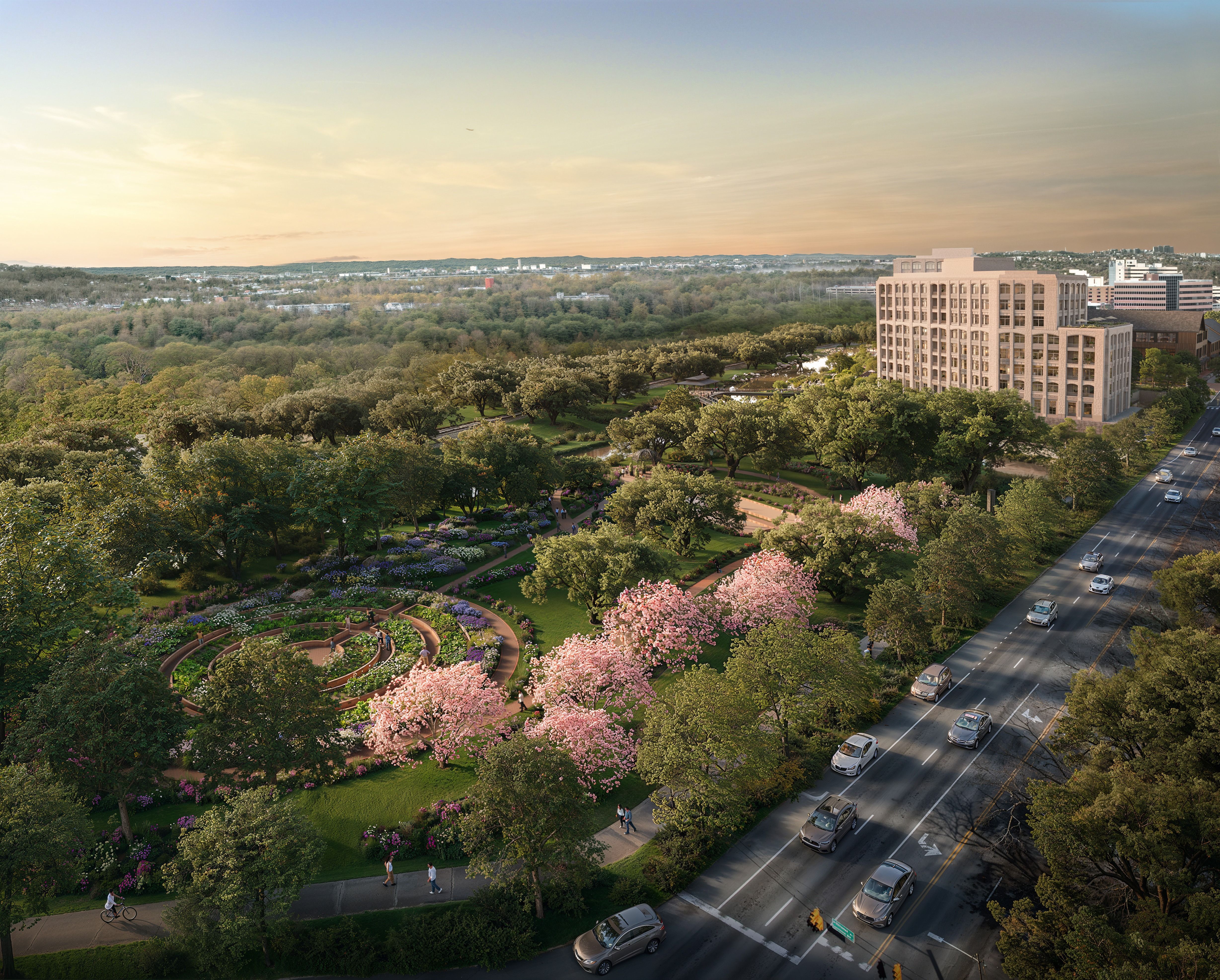 Aerial view of a city park with winding paths, blooming pink trees, and people walking and cycling. A multi-story building and cars on a road border the park under a clear sky.