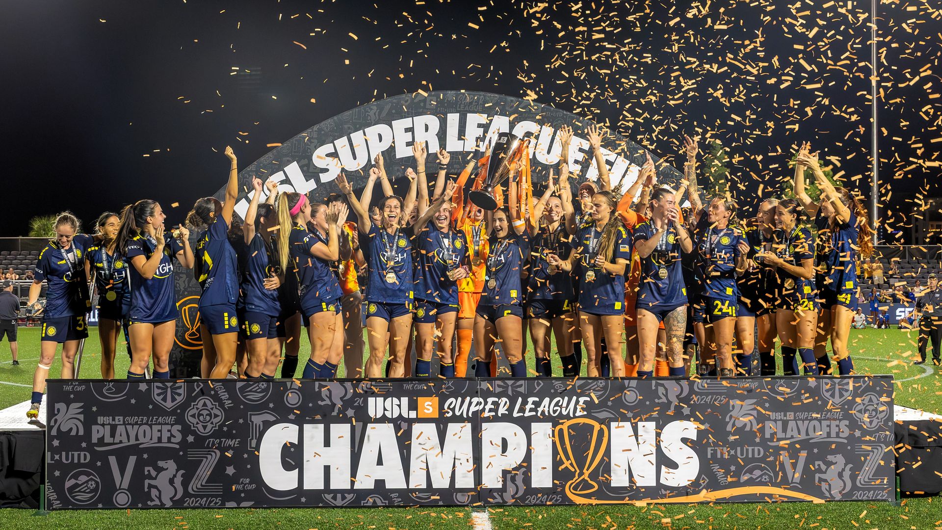 Soccer players cheer on a gray platform that says "CHAMPIONS" in white across the front as yellow confetti falls from the sky.