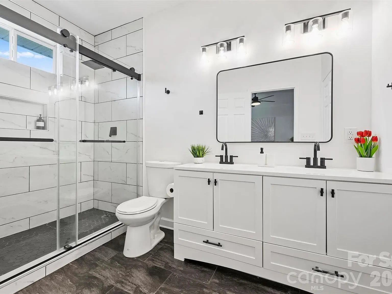 Bright modern bathroom with white cabinetry and countertops, black faucets, a large rounded mirror, glass sliding shower, white tile walls, dark floor, toilet, plus green plant and red tulips on the vanity.