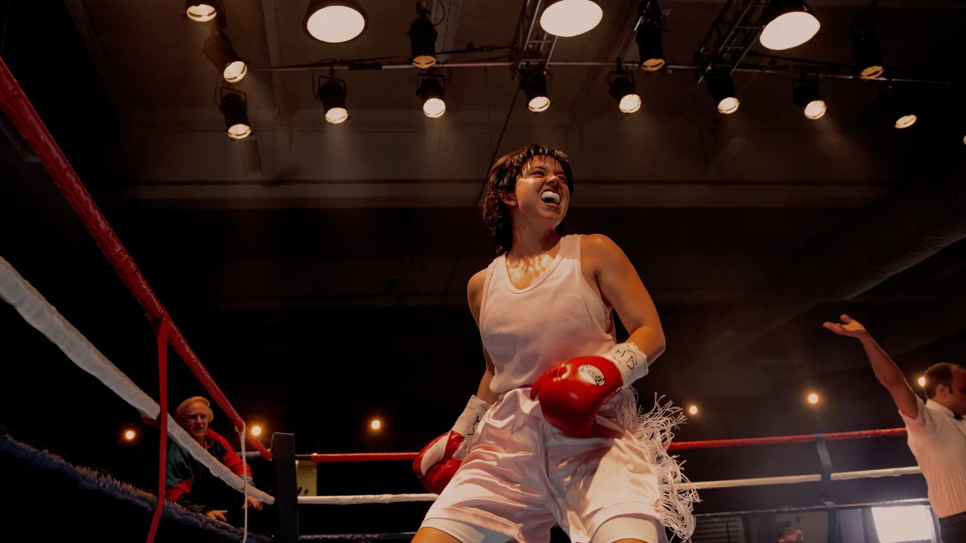 Female boxer in white outfit and red gloves celebrating a win inside a boxing ring under bright overhead lights, with a coach and referee in the background.