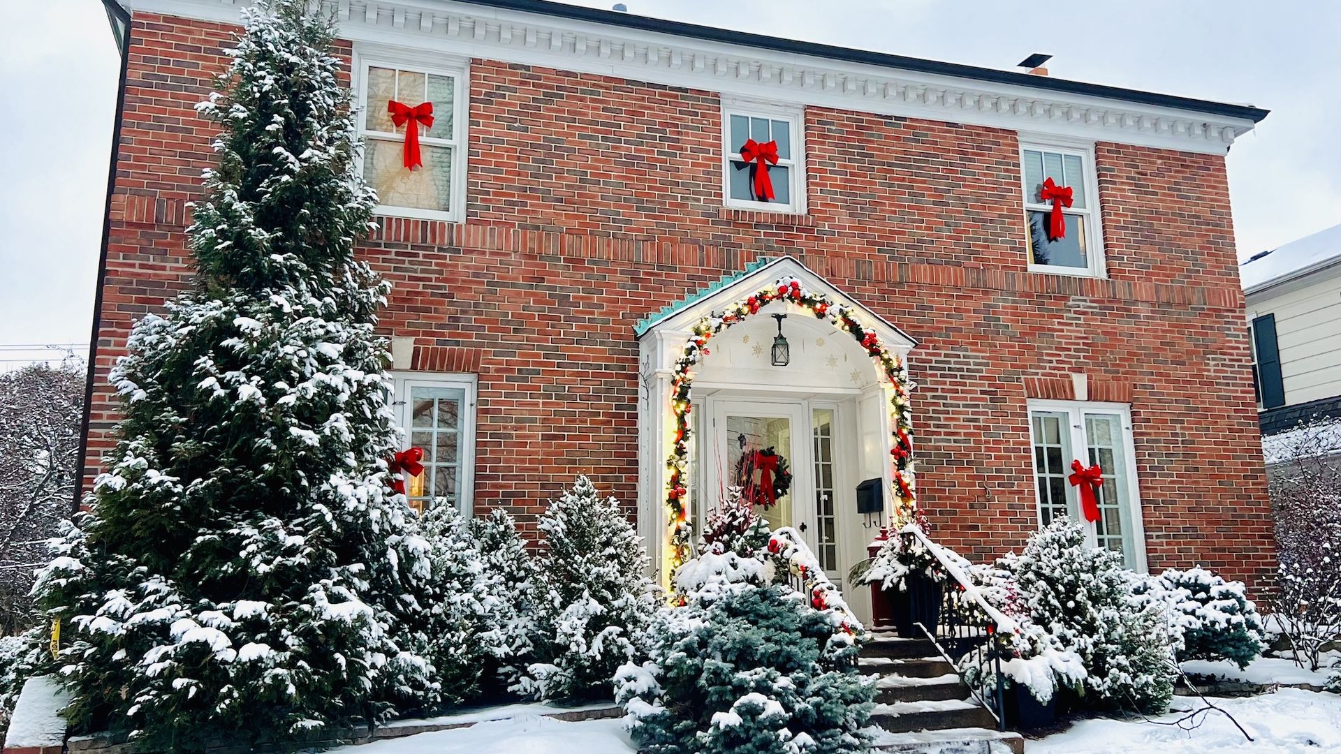 Snow-covered brick house decorated for Christmas with red bows on windows and railing, a wreath on the door, and garland with lights around a white porch entrance.