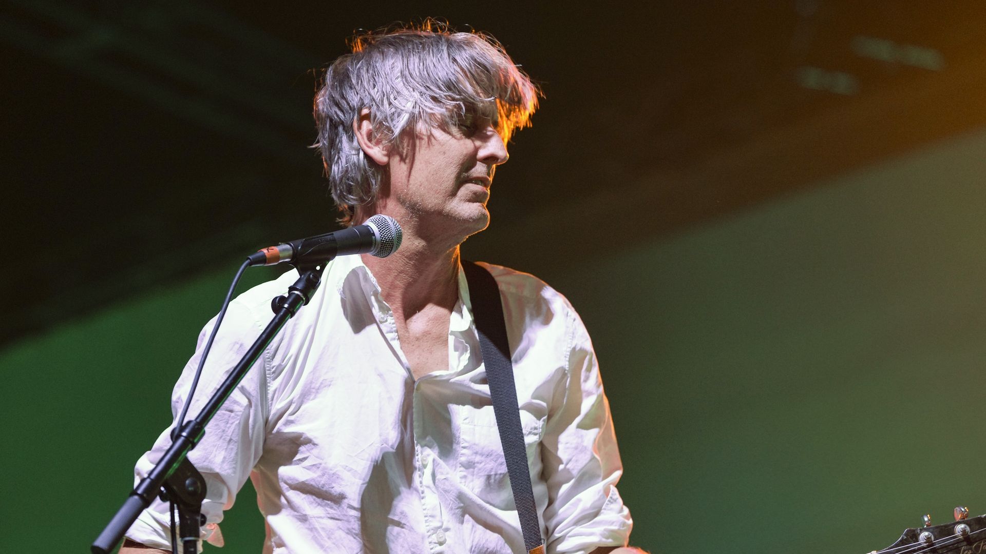 Man with gray hair in a white rolled-up shirt playing a brown Les Paul Deluxe guitar and singing into a microphone on stage under warm lighting.