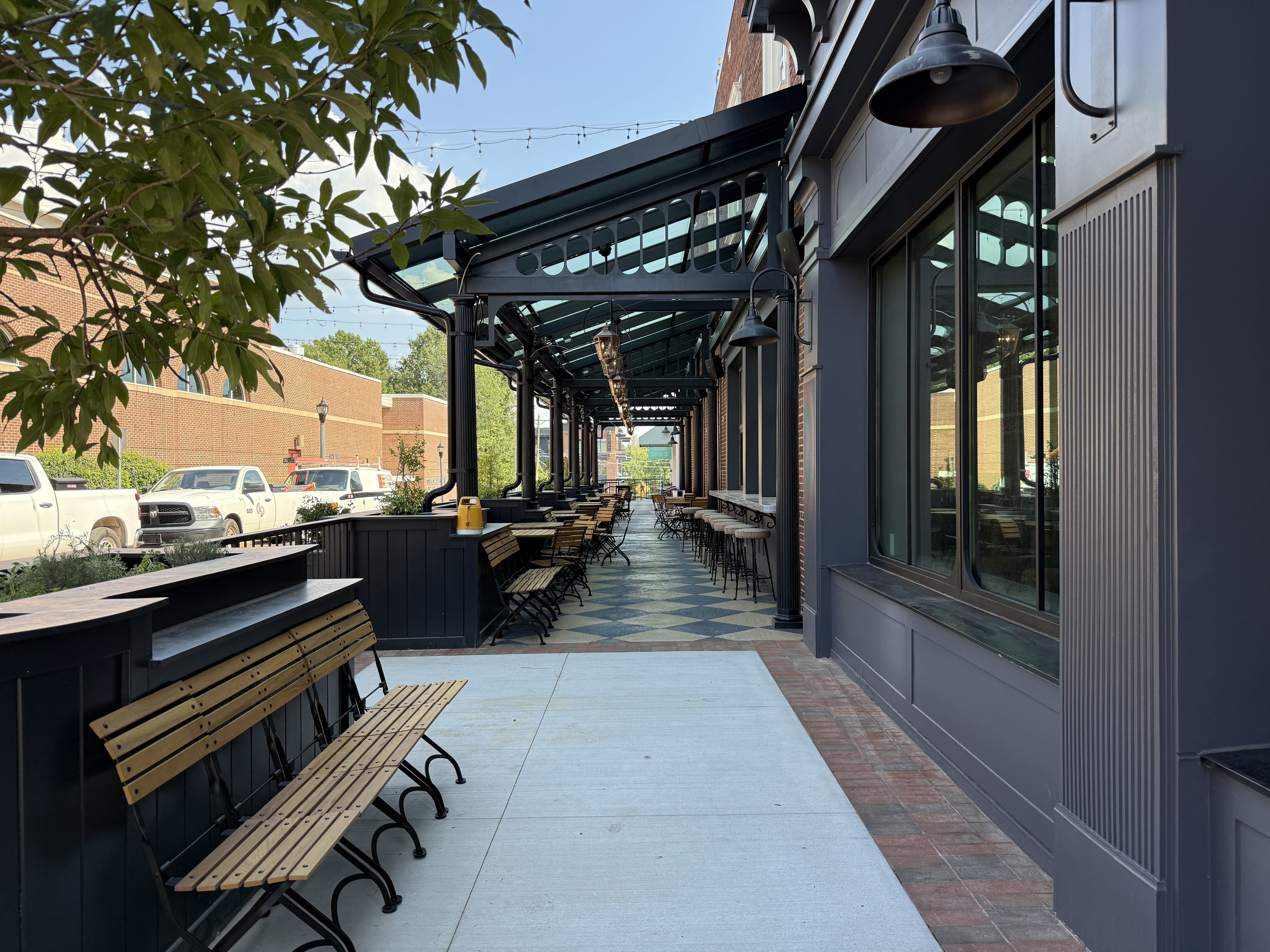 Outdoor walkway with black metal pergola, string lights, and rows of wooden benches beside a dark blue storefront; brick buildings, trees, and parked cars in the background.