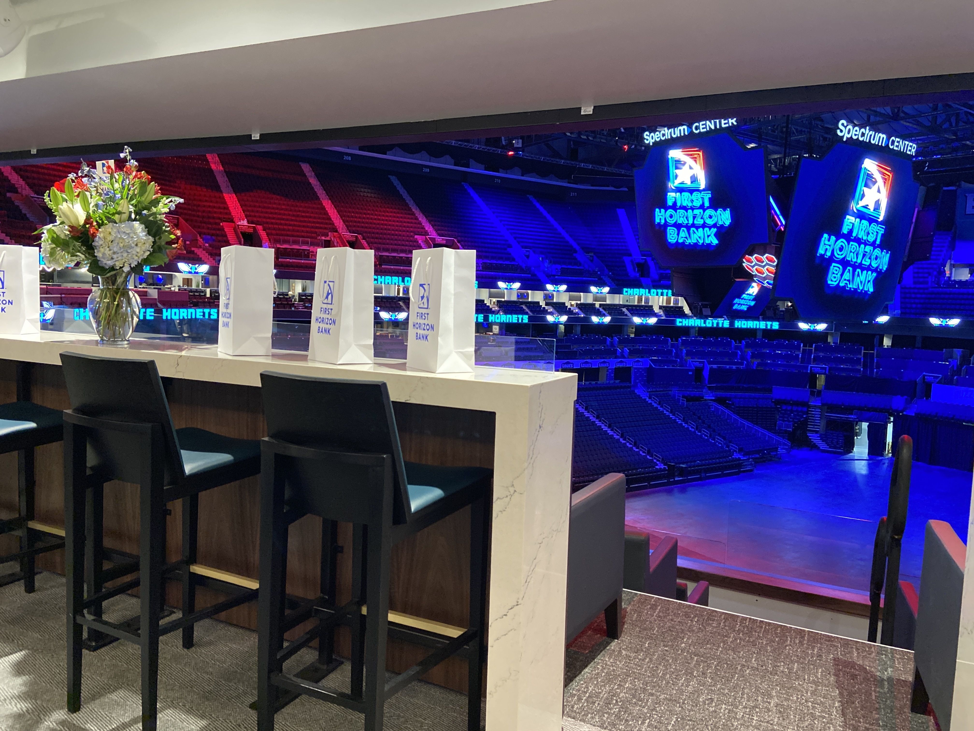  A VIP suite inside Spectrum Center, overlooking the arena floor. The suite has a high-top counter with black barstools. A bouquet of fresh flowers sits on the counter as decoration. The background shows the seating
