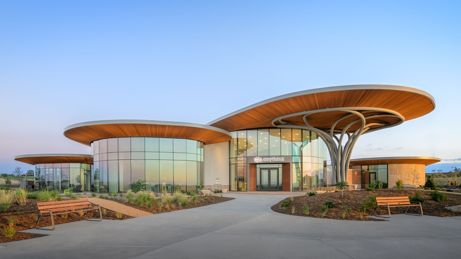 A futuristic public building with large curved wooden canopies supported by gray metal arches, a glass facade, and a surrounding landscaped plaza with benches under a clear blue sky.