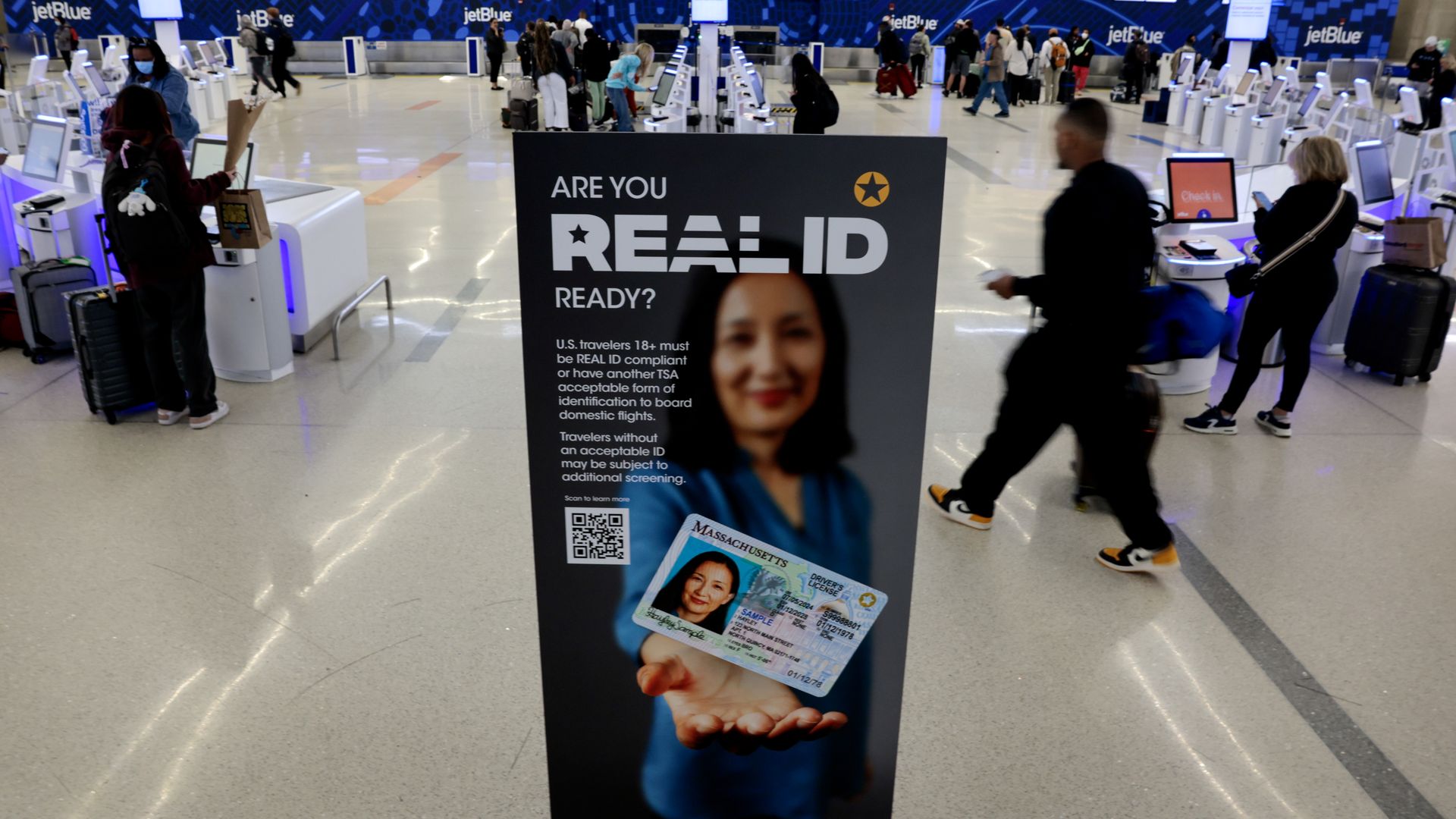 Airport check-in area with JetBlue signage and self-service kiosks, featuring a prominent sign asking "Are you REAL ID ready?" showing a woman holding a Massachusetts driver's license.