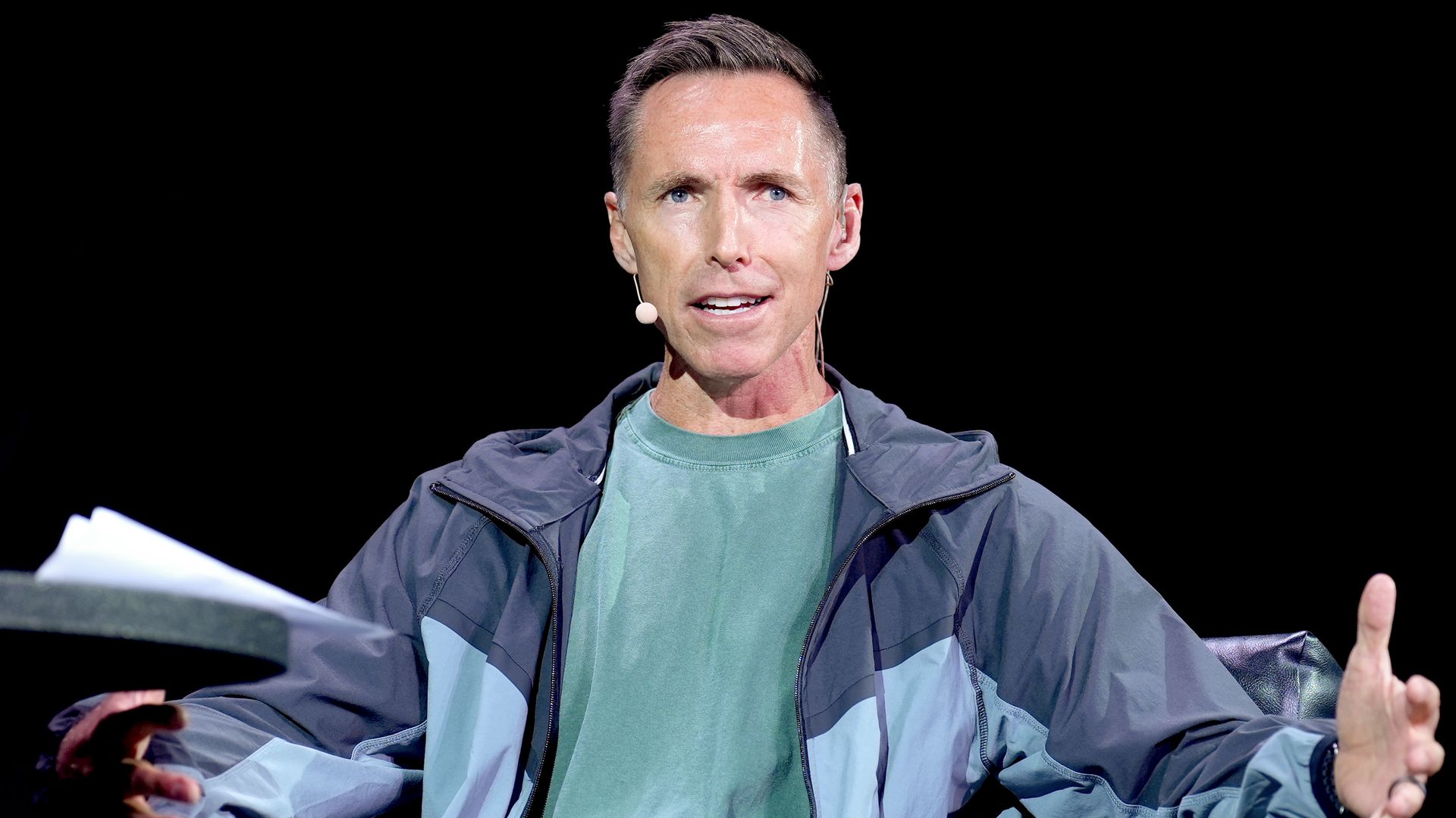 Steve Nash, wearing a blue jacket over a green t-shirt with a small microphone extending from his ear to his mouth sits while holding his hands out to his side. 