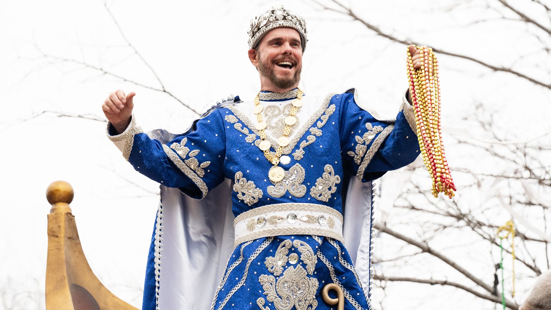 Kevin Dillon, dressed as Renaissance-era royalty, holds Mardi gras beads and smiles at a crowd from atop a float.