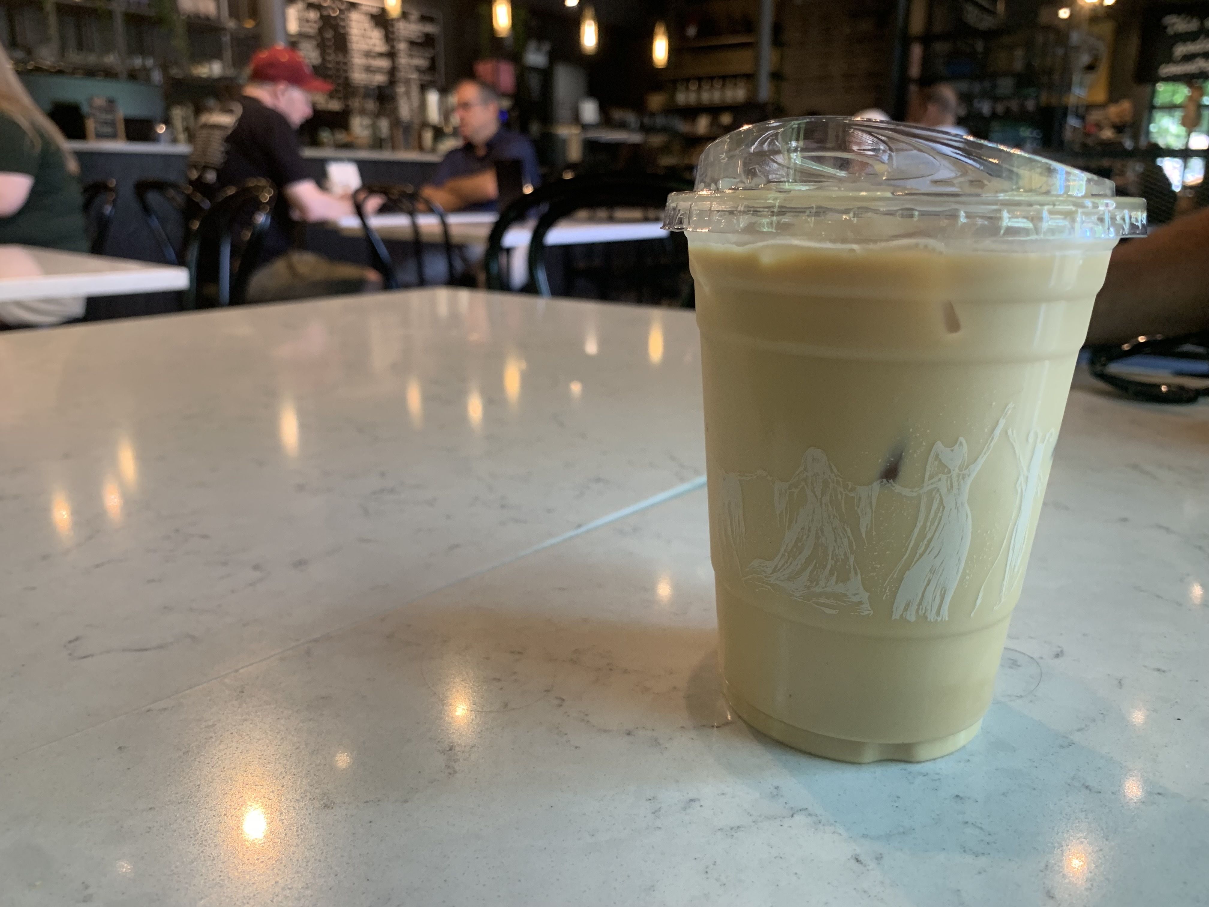 Close-up of a plastic cup with a clear lid on a white marble counter, holding a pale green iced drink. In the blurred background, a cafe with patrons, a bar, and a chalkboard menu.