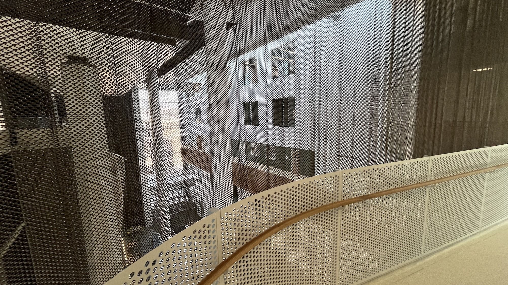 Interior atrium viewed from a walkway: a gray perforated metal mesh screen in the foreground, with a curved white railing and a wooden handrail. Surrounding building facades with windows.