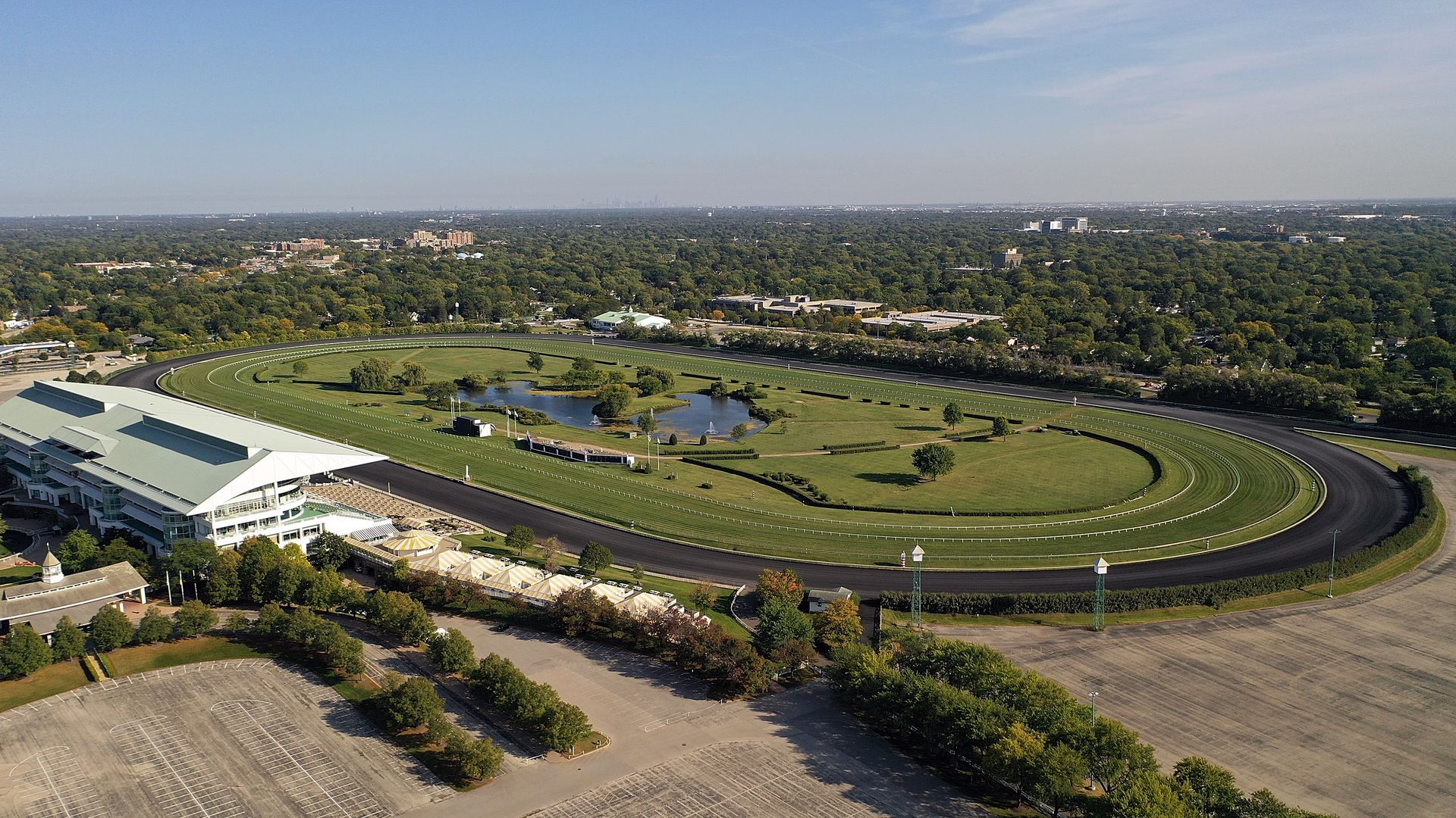Aerial view of a large horse racing track with green grass, a pond in the center, surrounding grandstands, parking lots, and a densely treed urban area in the background under a clear sky.
