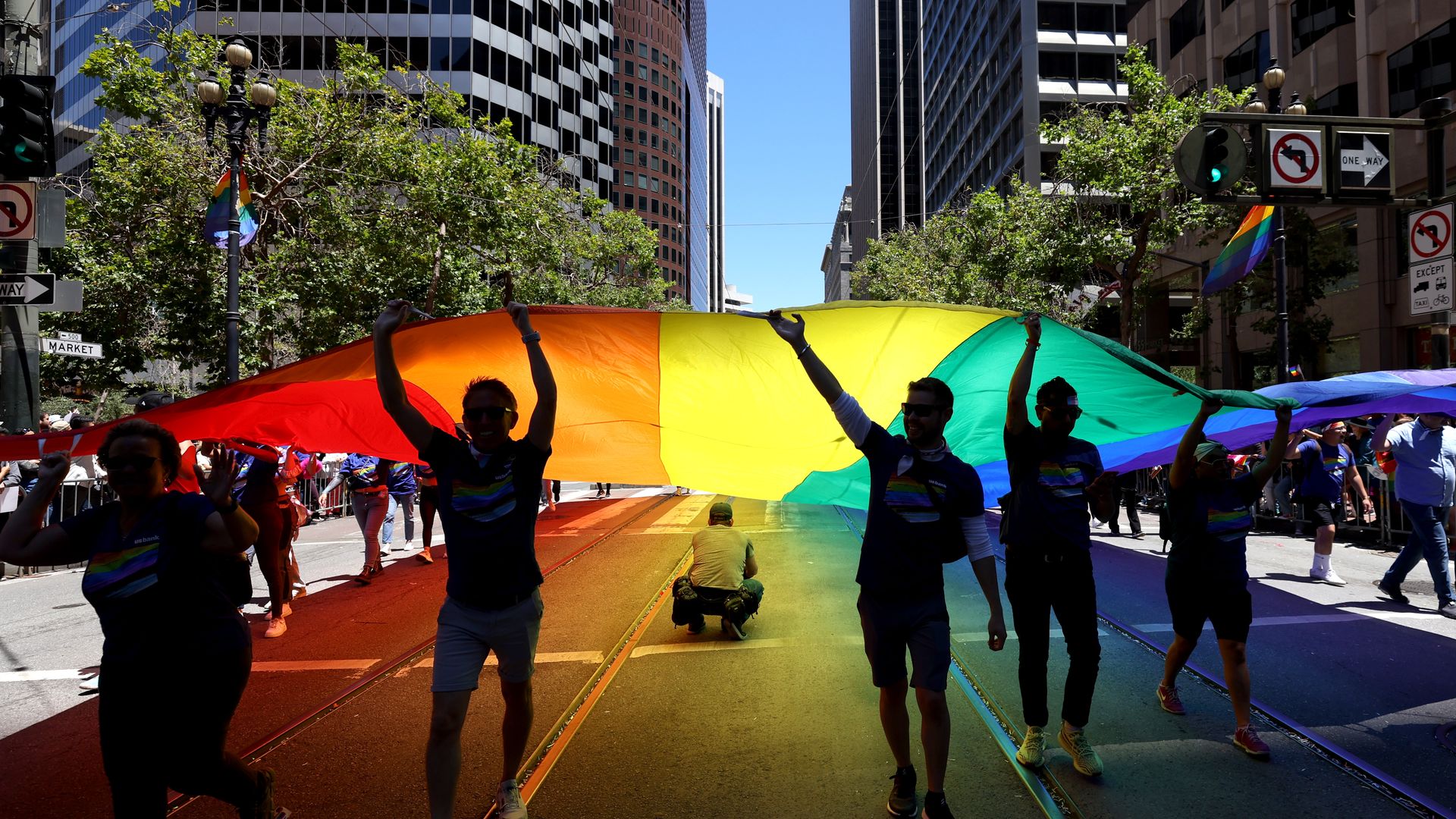 People participate in a city Pride parade, lifting a huge rainbow banner over tram tracks; rainbow colors stretch along the road amid tall buildings and green trees.