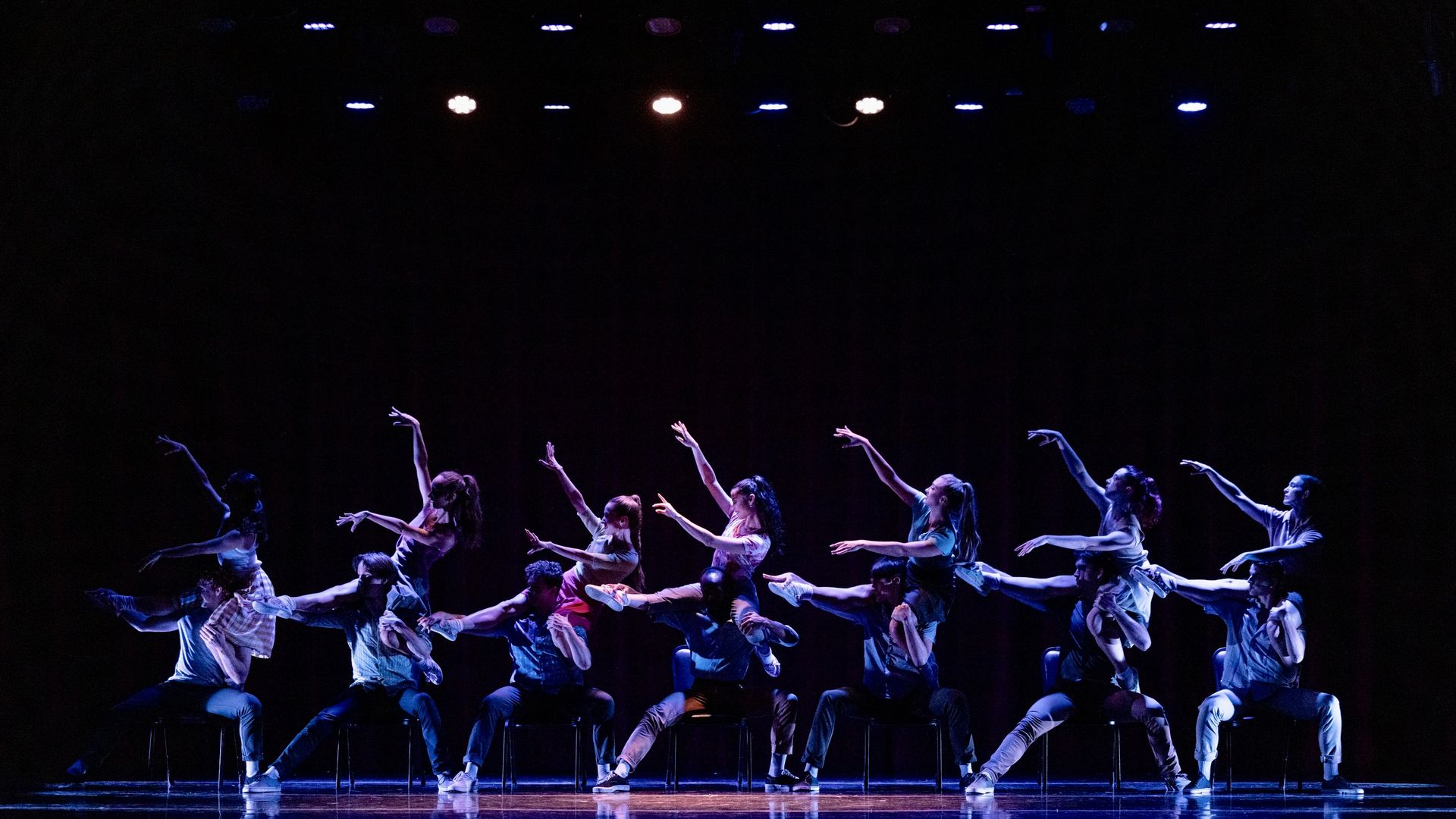 Group of performers on stage, six men seated on chairs lifting six women in dynamic ballet poses, under purple and blue stage lights against a black background.