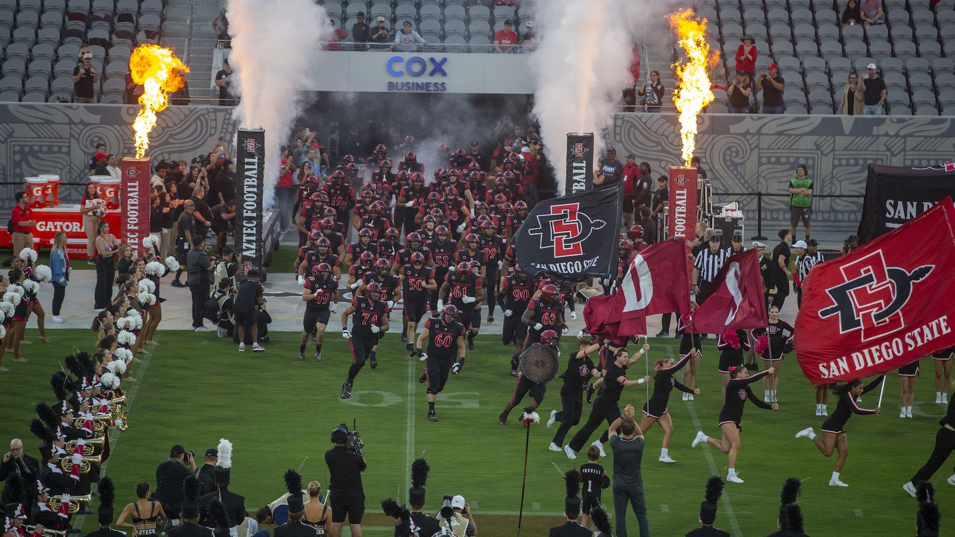 San Diego State football players in black uniforms run onto the field through smoke and flames, cheered by cheerleaders with red flags and a marching band in black uniforms.