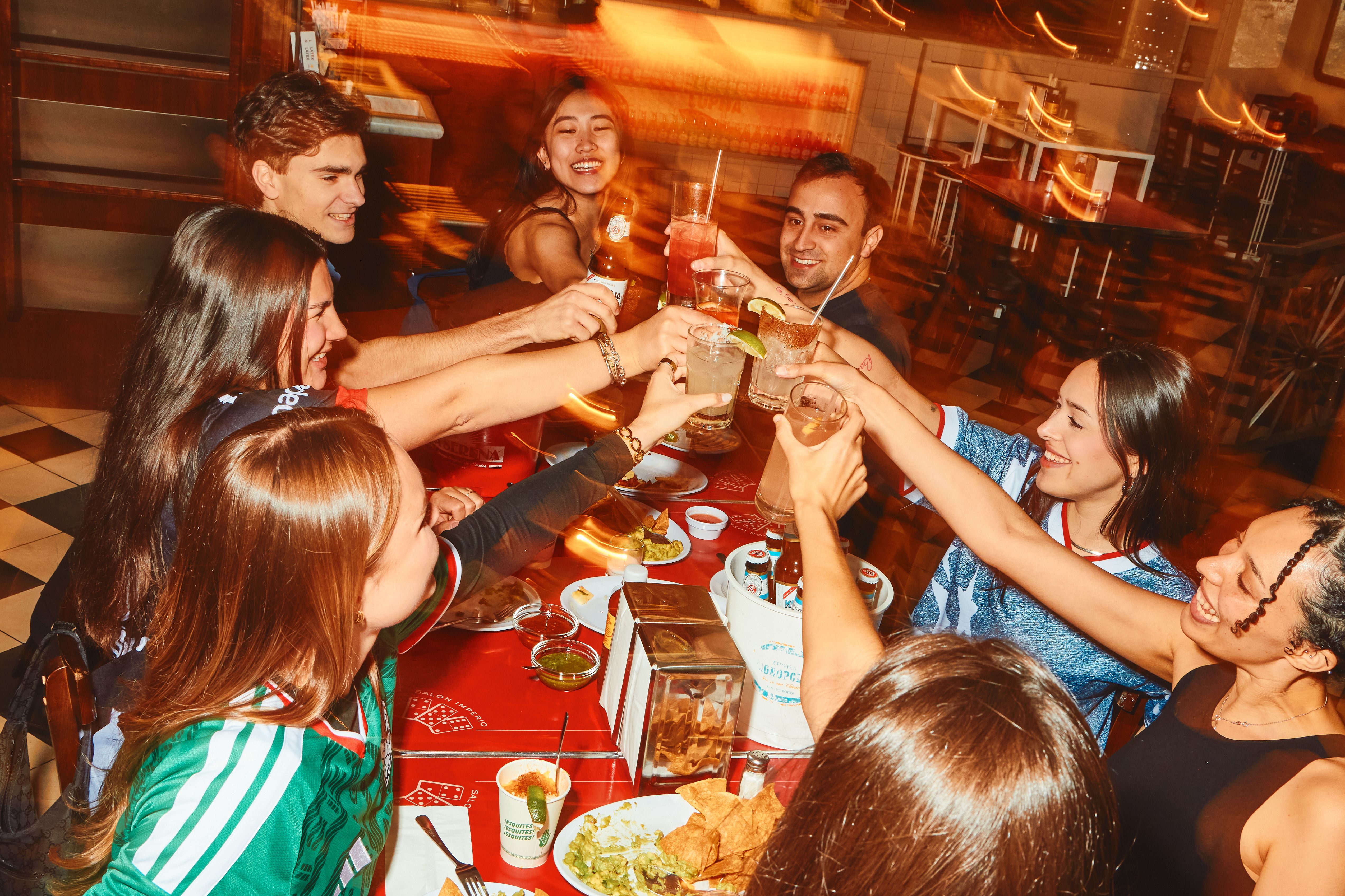 A diverse group of friends at a crowded, warm orange-lit restaurant raise glasses in a toast, smiling as beer bottles and colorful cocktails clink above a red-checkered table.