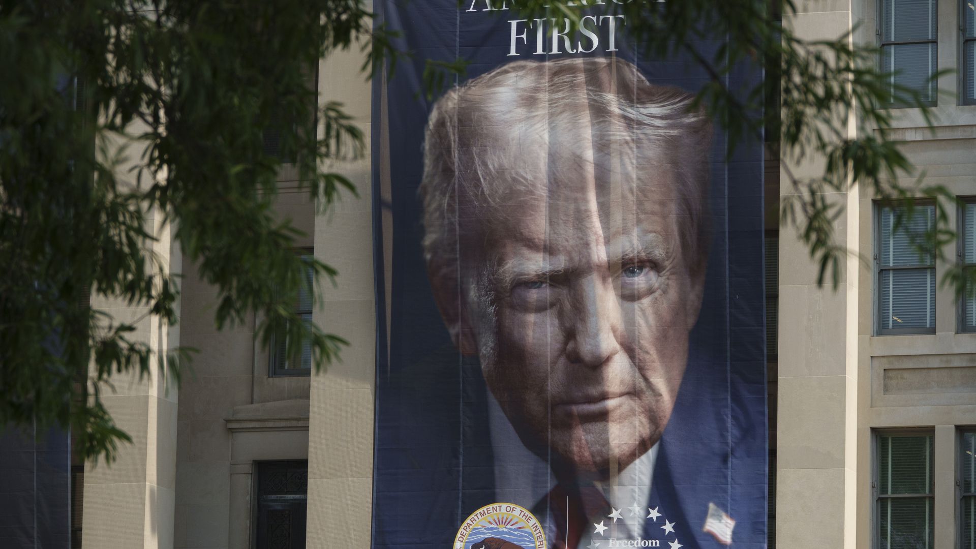 A large banner of President Trump's face and shoulders against a blue backdrop hangs on the front of a tan government building. In the photo, he holds a straight face and is wearing a dark blue suit, a white collared shirt, a red tie and an American flag pin.