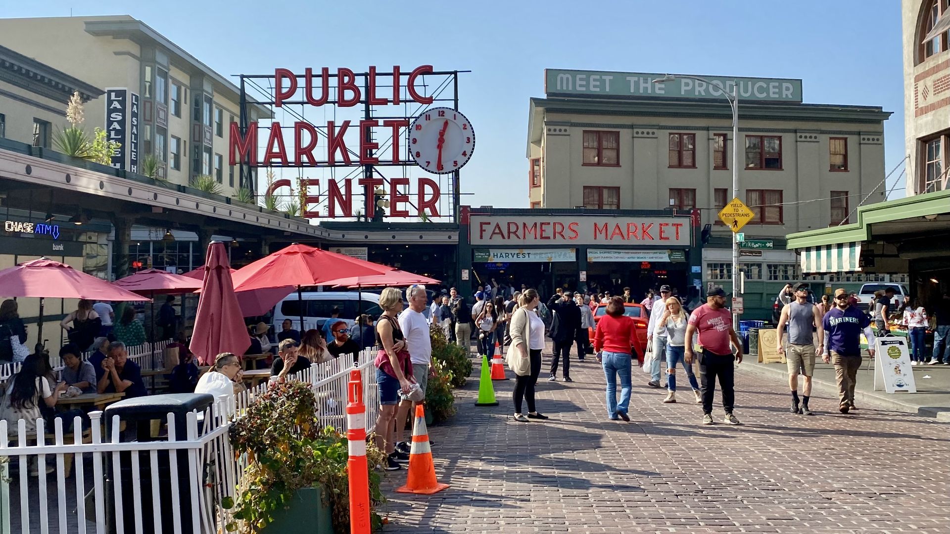 People walk along a brick street in front of the Pike Place Market neon sign that says "Public Market Center" and an awning that says "farmers market."