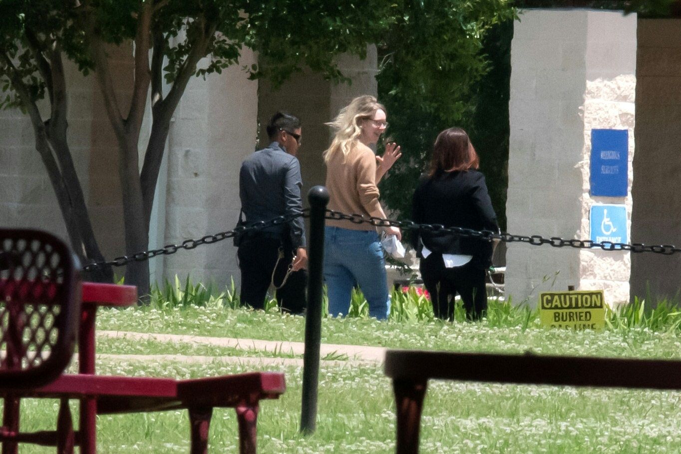 Three people walking outside near a stone building, one person with blonde hair waving, trees and greenery around, red picnic table in foreground, caution sign about buried gas line.