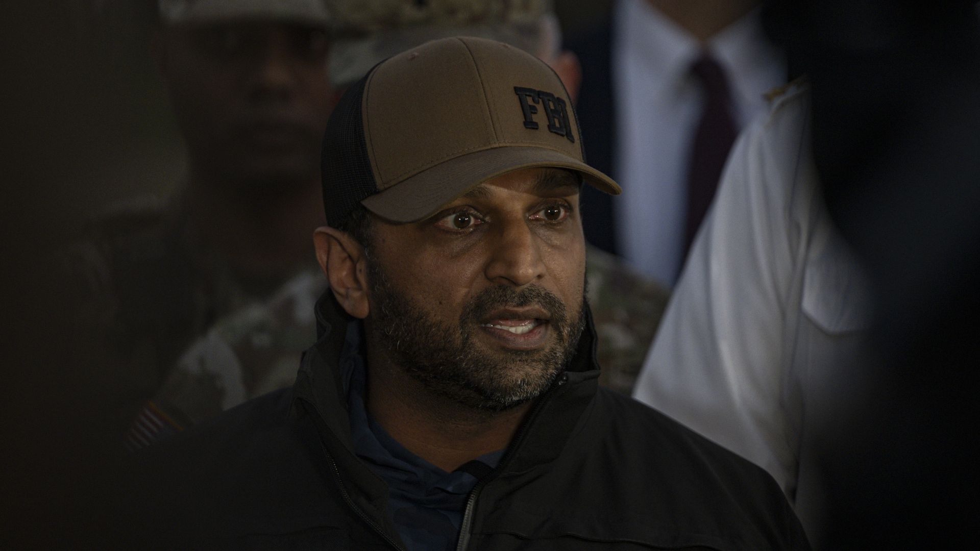 Kath Patel, wearing a black and brown FBI hat and a black windbreaker, standing in front of several other people wearing military fatigues, police uniforms and suits.