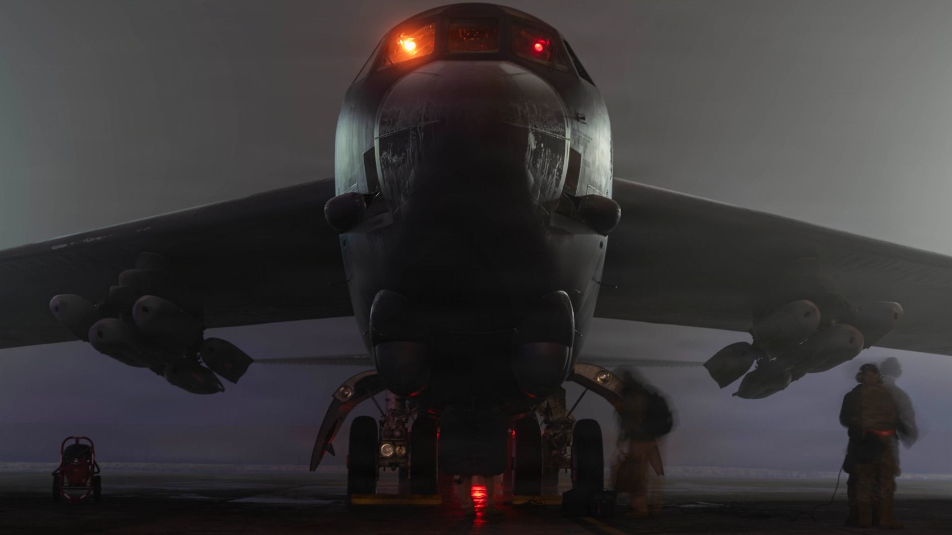 Front view of a large military aircraft with landing gear down, wing-mounted pods, illuminated orange and red lights, and two figures standing near the right wing in foggy, low-light conditions.