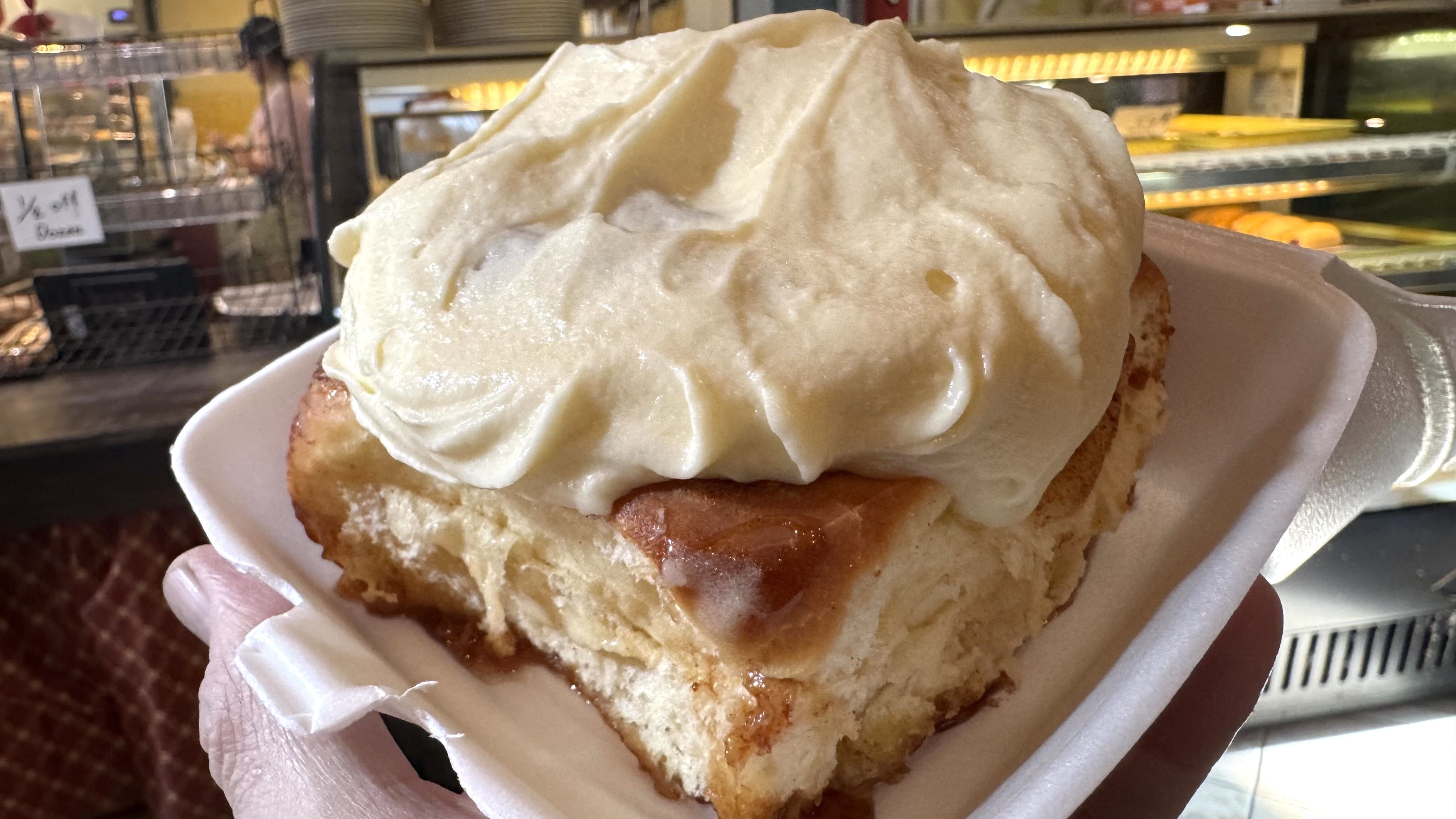 Close-up of a large cinnamon roll with thick white frosting served on a white foam tray, held in a hand inside a bakery setting.