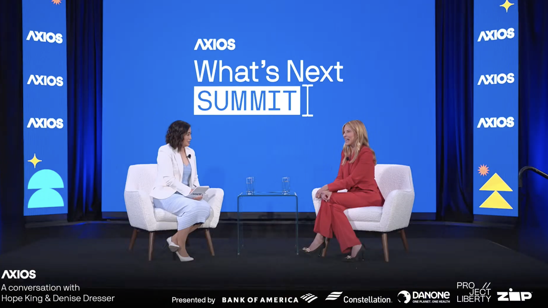 Two people sit in chairs during an interview on a stage with a backdrop saying Axios What's Next Summit