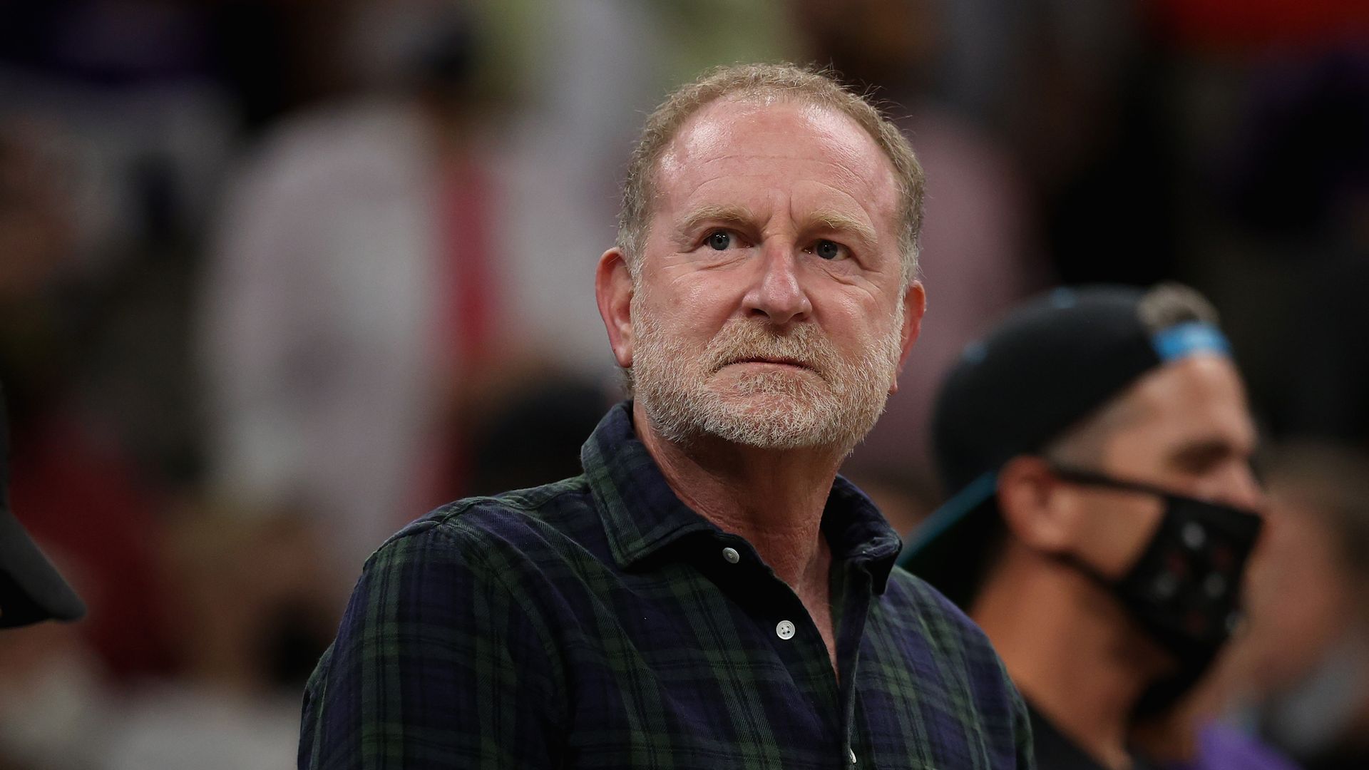 A man wearing a button-up shirt with a green checkered pattern over dark blue looks ahead with blurry images of an arena crowd in the background; a man in a black face mask and a black baseball cap turned backwards is visible behind him.