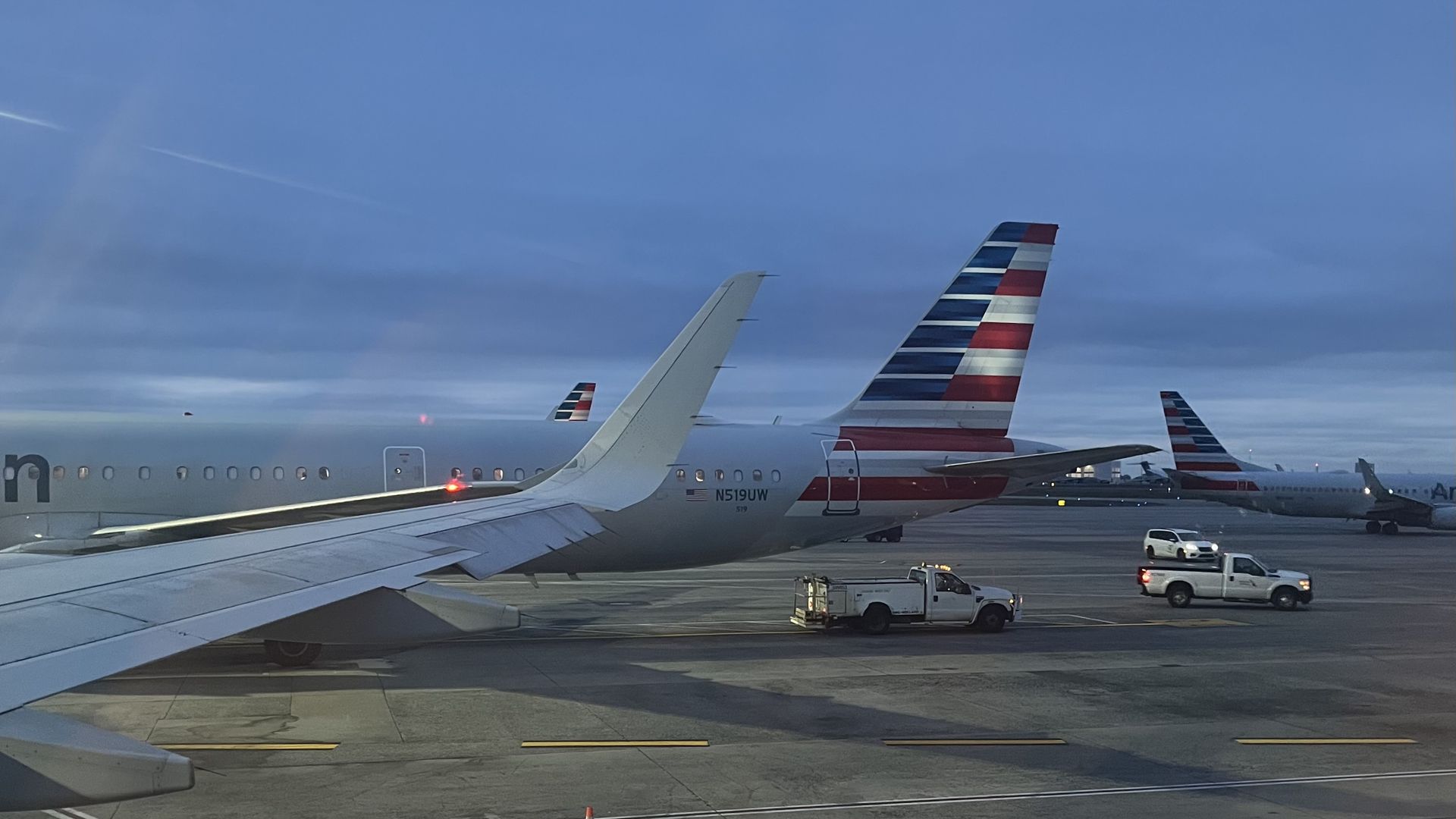Ground view of several US airliners on the tarmac, dominated by American Airlines tails with red, white, and blue stripes. Ground vehicles and orange cones near the planes under a blue sky.