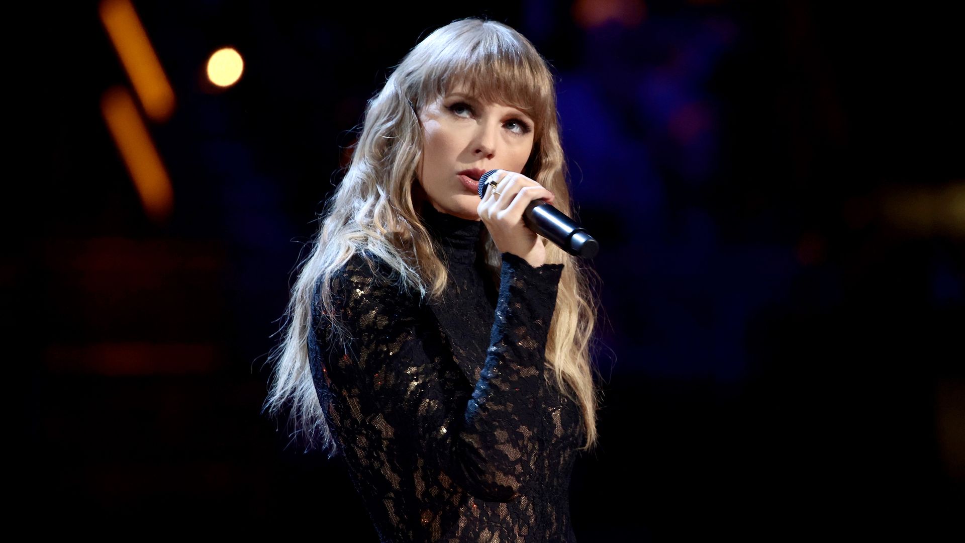 Taylor Swift sings into a microphone while standing on stage in a black lace outfit. 