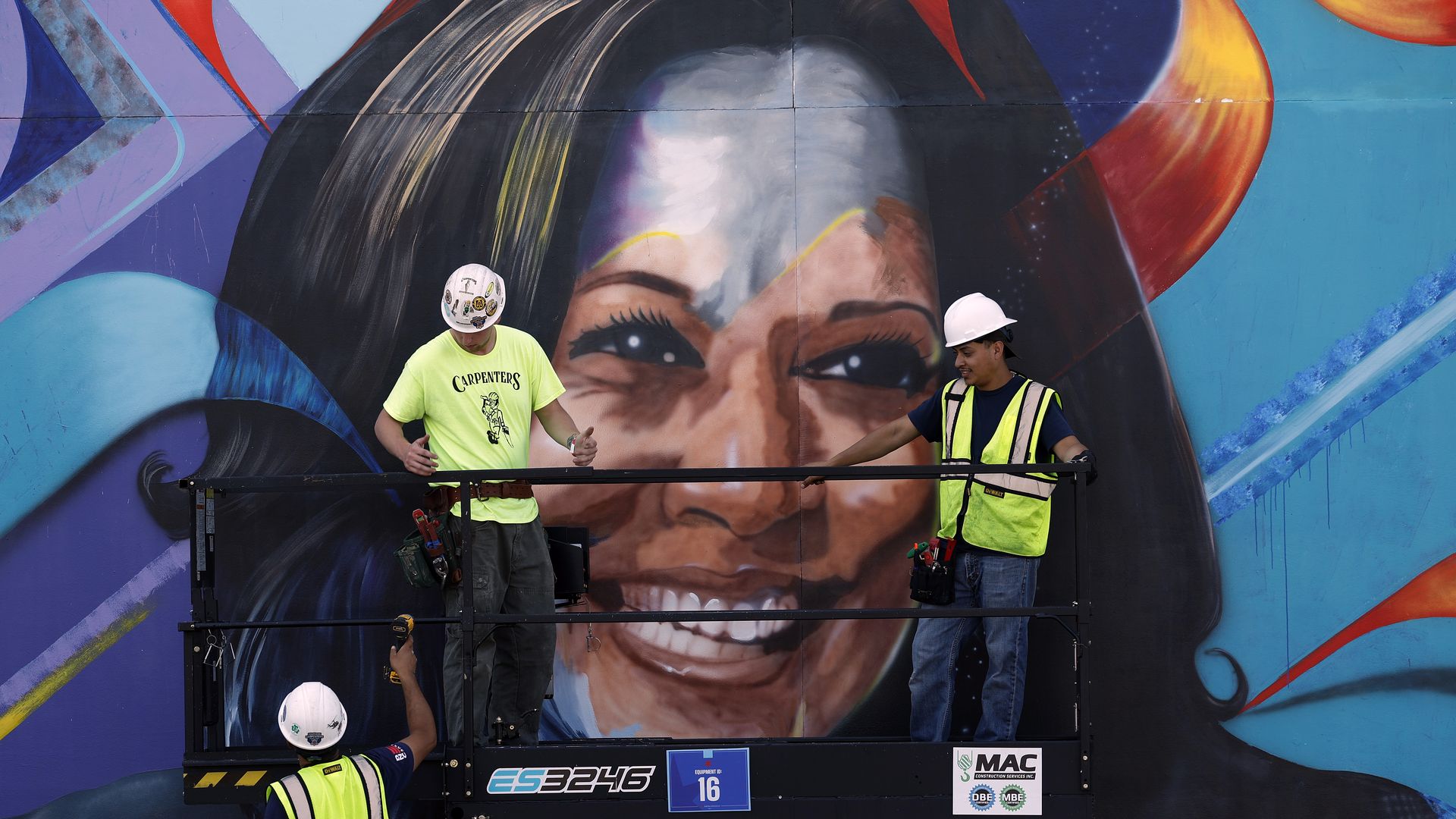 Three people in hard hats work on a large, colorful mural