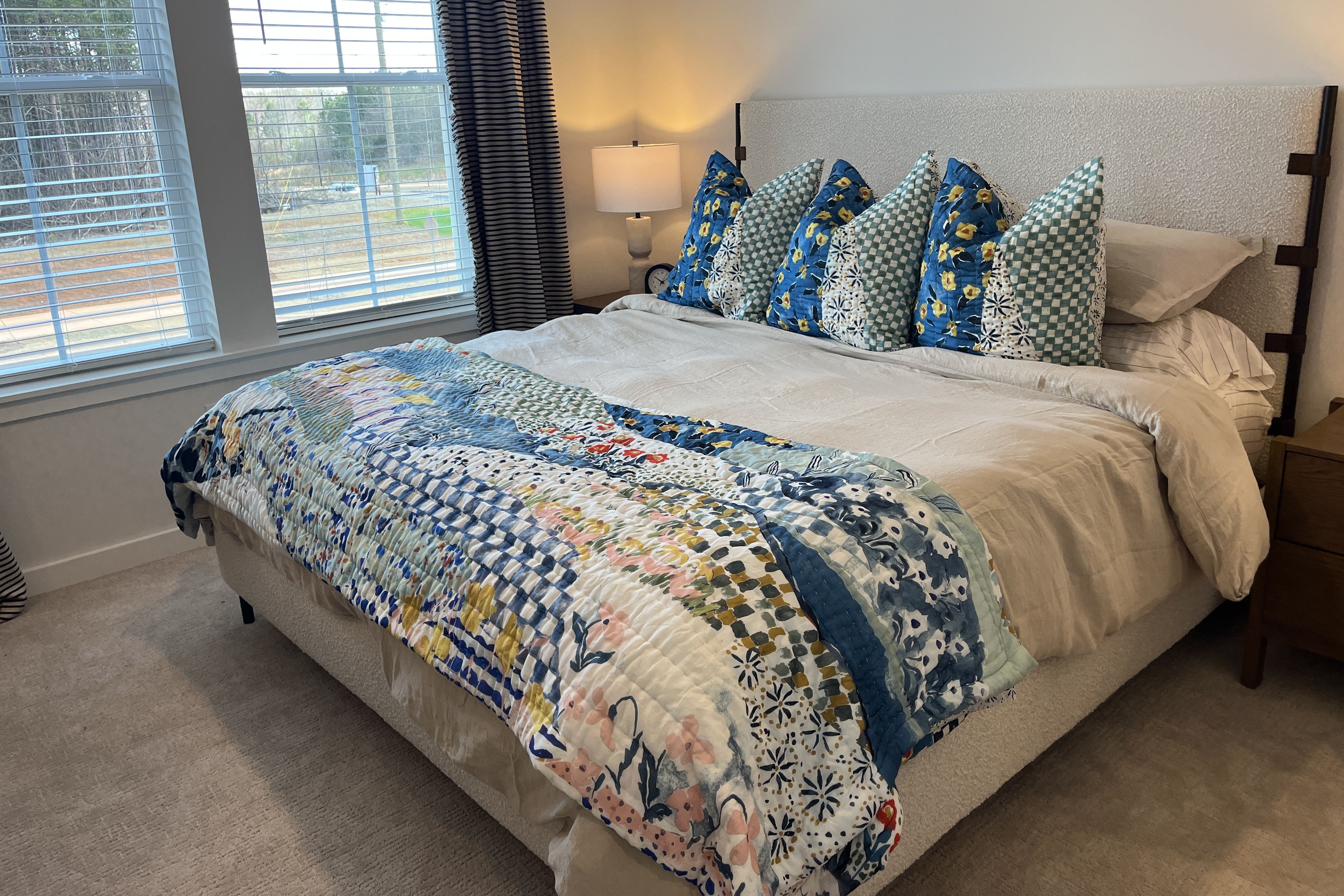 Cozy bedroom with a large bed, white sheets, and a colorful patchwork quilt at the foot. Blue and teal pillows sit against a light, textured headboard; lamp on left nightstand, window blinds visible.