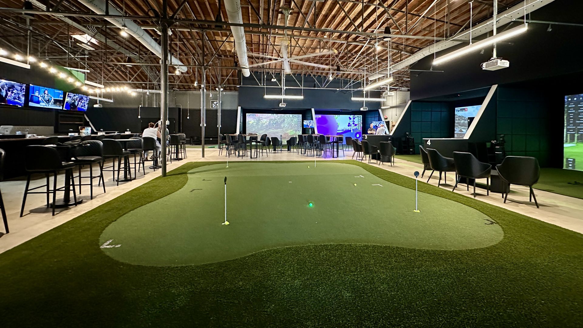 Indoor golf simulation center with putting green in foreground, tall black chairs and tables on left, multiple golf simulators with screens on right, and exposed wooden ceiling with lights.