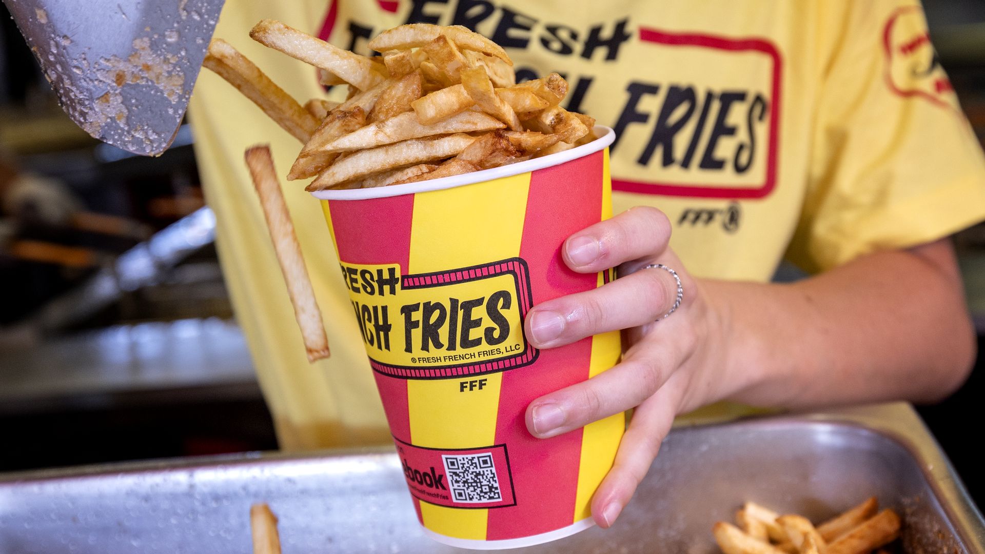 Person in a yellow shirt holds a red-and-yellow striped cup of French fries; a fry sticks out of the cup while more fries spill onto a metal tray.