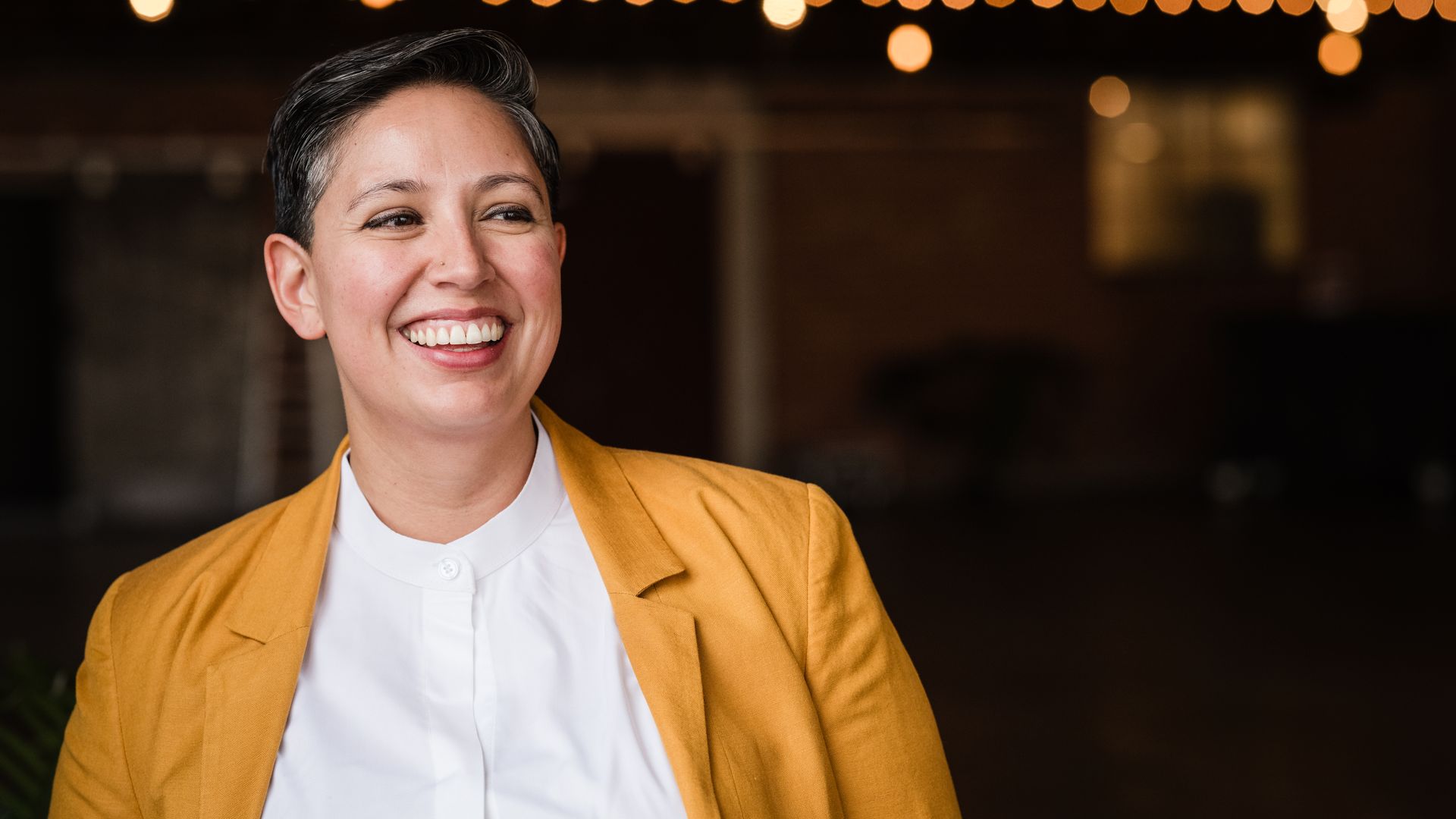 Smiling person with short dark hair wearing a mustard yellow jacket and white shirt, with blurred string lights in the dark background.