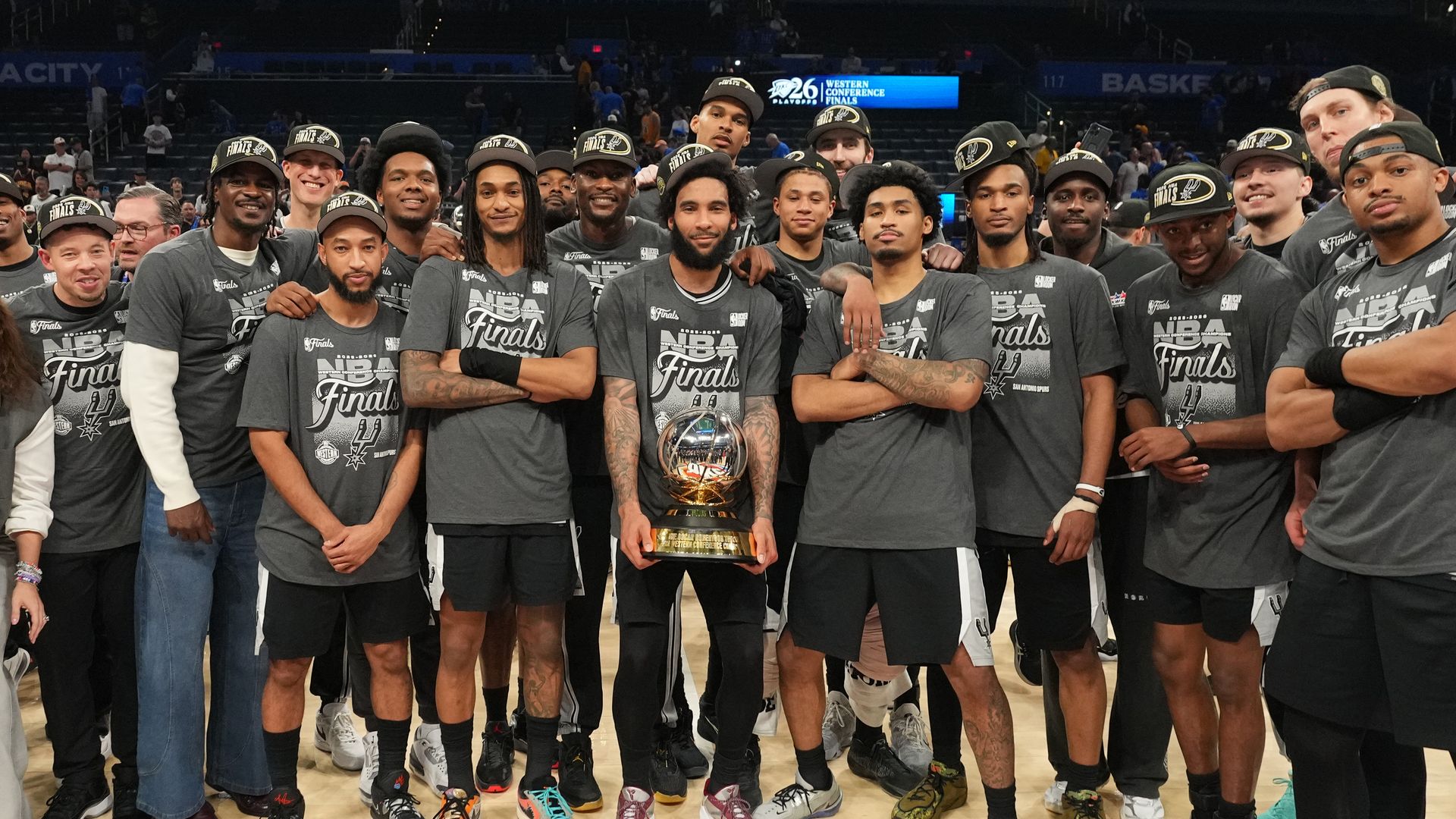 Group of basketball players in gray Finals shirts and caps pose together on a court, centered around a shiny trophy held by a bearded man. Arena lights and spectators in the background.