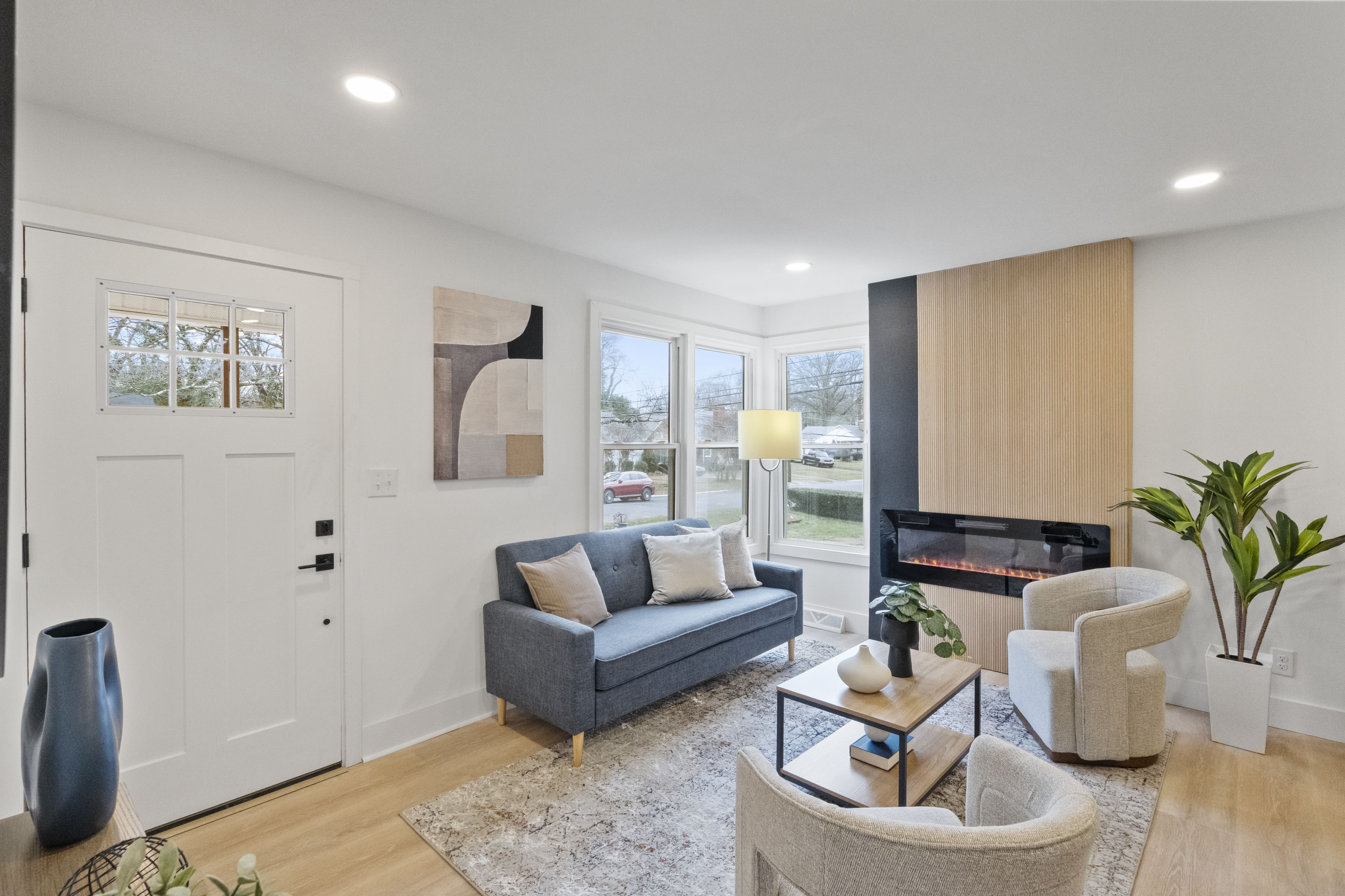 Modern living room with wooden floor, blue sofa, beige armchairs, light wood coffee table, electric fireplace with wood panel background, and large windows showing outdoors.