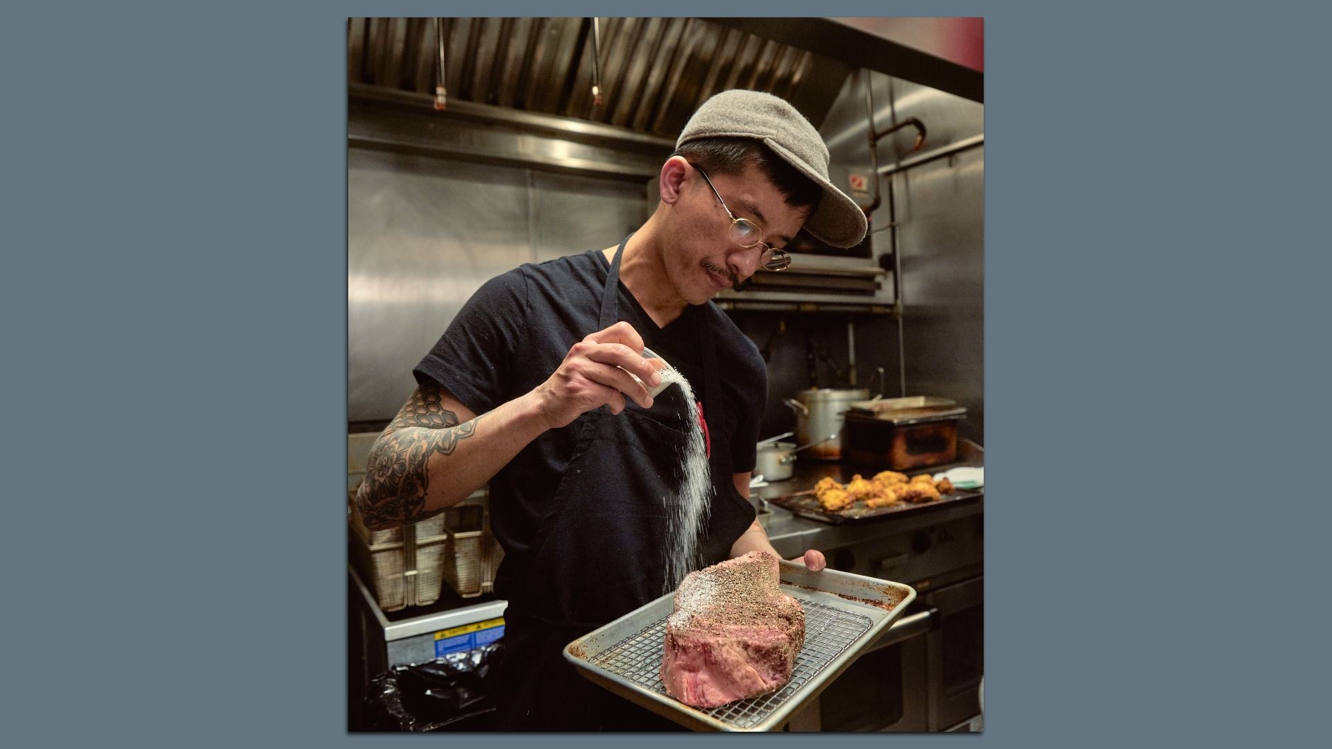 Man with a tattoo on his arm wearing glasses, a gray cap, and black apron seasoning a large piece of raw meat in a professional kitchen with cooked chicken in the background.