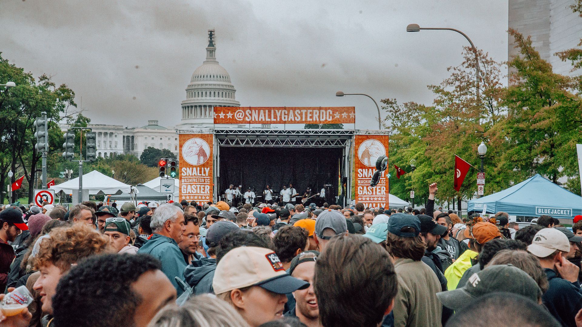 A crowded outdoor festival with a large orange stage, banners reading "SNALLYGASTER DC"; the U.S. Capitol dome is visible in the background under a gray sky.