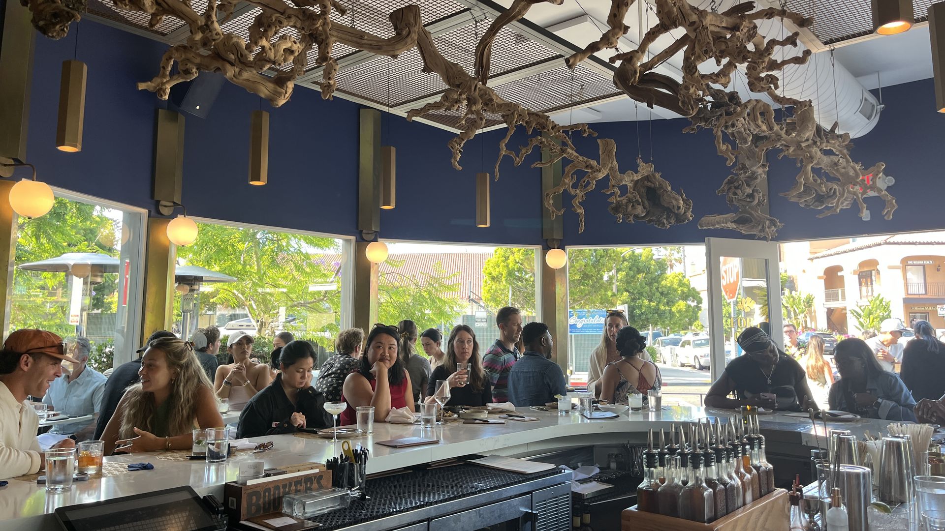 Busy bar area with people sitting along a white curved counter, large windows showing green trees outside, and twisted wooden decor hanging from the blue ceiling.