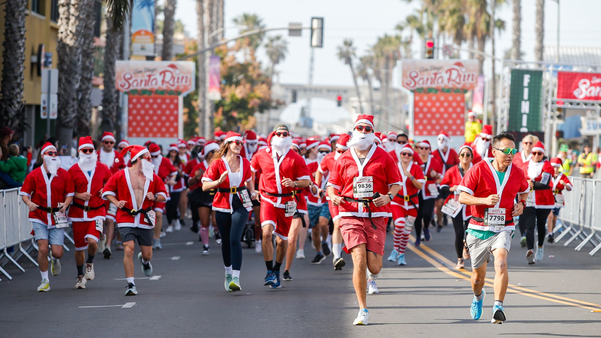 A large group of runners dressed in red and white Santa Claus outfits participating in the San Diego Santa Run on a palm tree-lined street during the daytime.