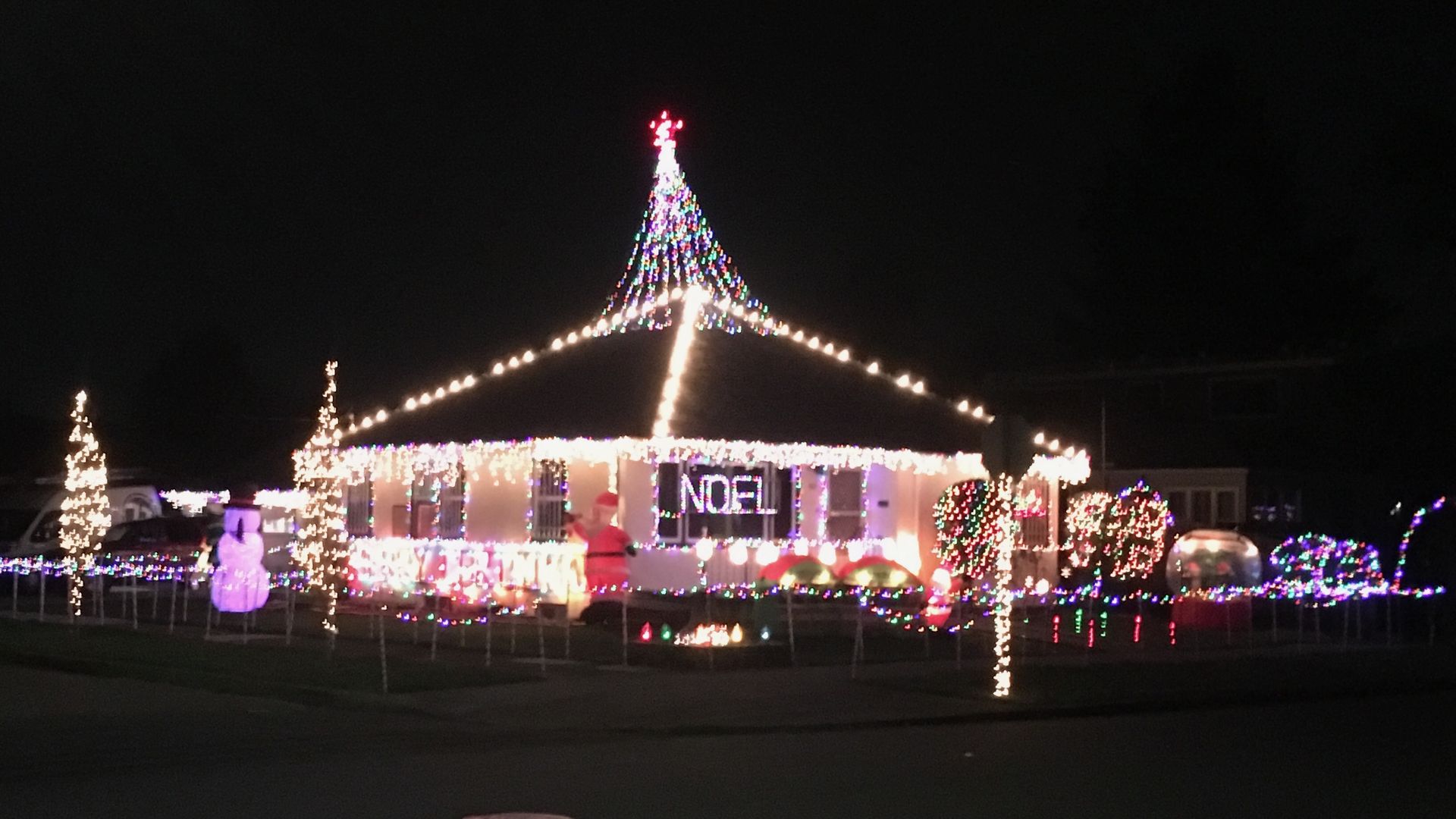 A single story house with extensive holiday lights, including a cone of lights on the roof representing a Christmas tree, and the word Noel in white lights in the front window.