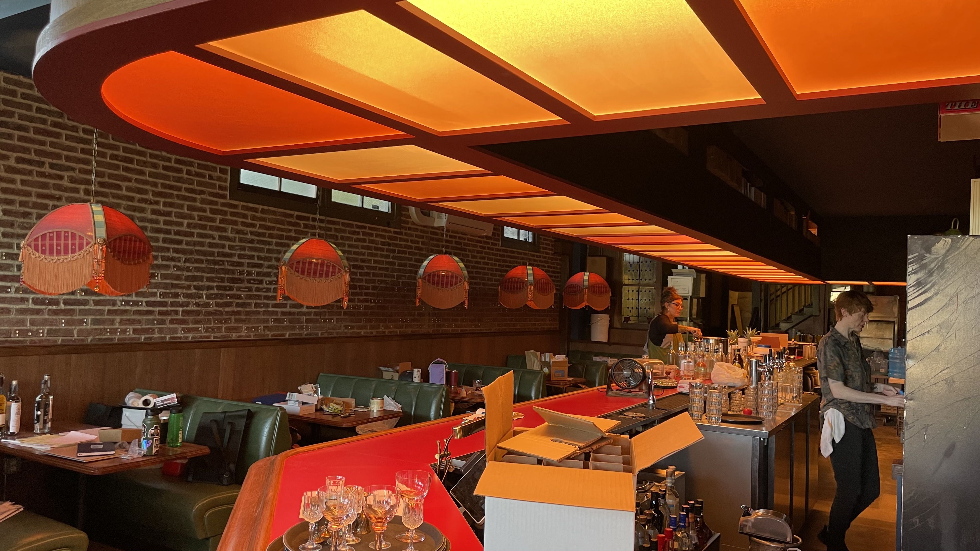 Interior of a cozy bar with glowing orange ceiling lights, red bar counter, green seats, vintage red lamps, and two staff members working behind the bar.
