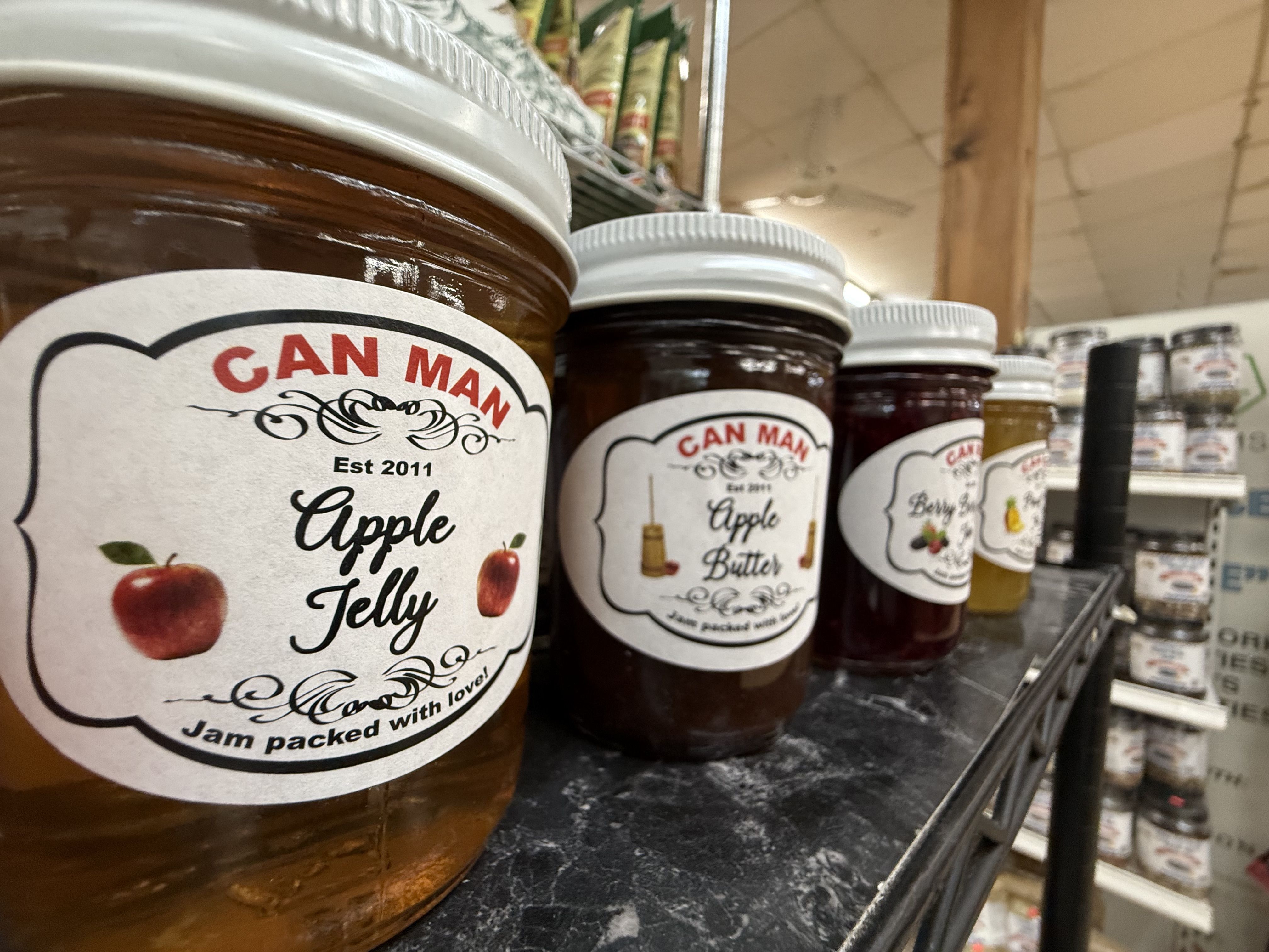 Close-up of Can Man jam jars on a store shelf, labels showing "Apple Jelly" with red apples, white lids, amber jam, and a blurred grocery aisle in the background.