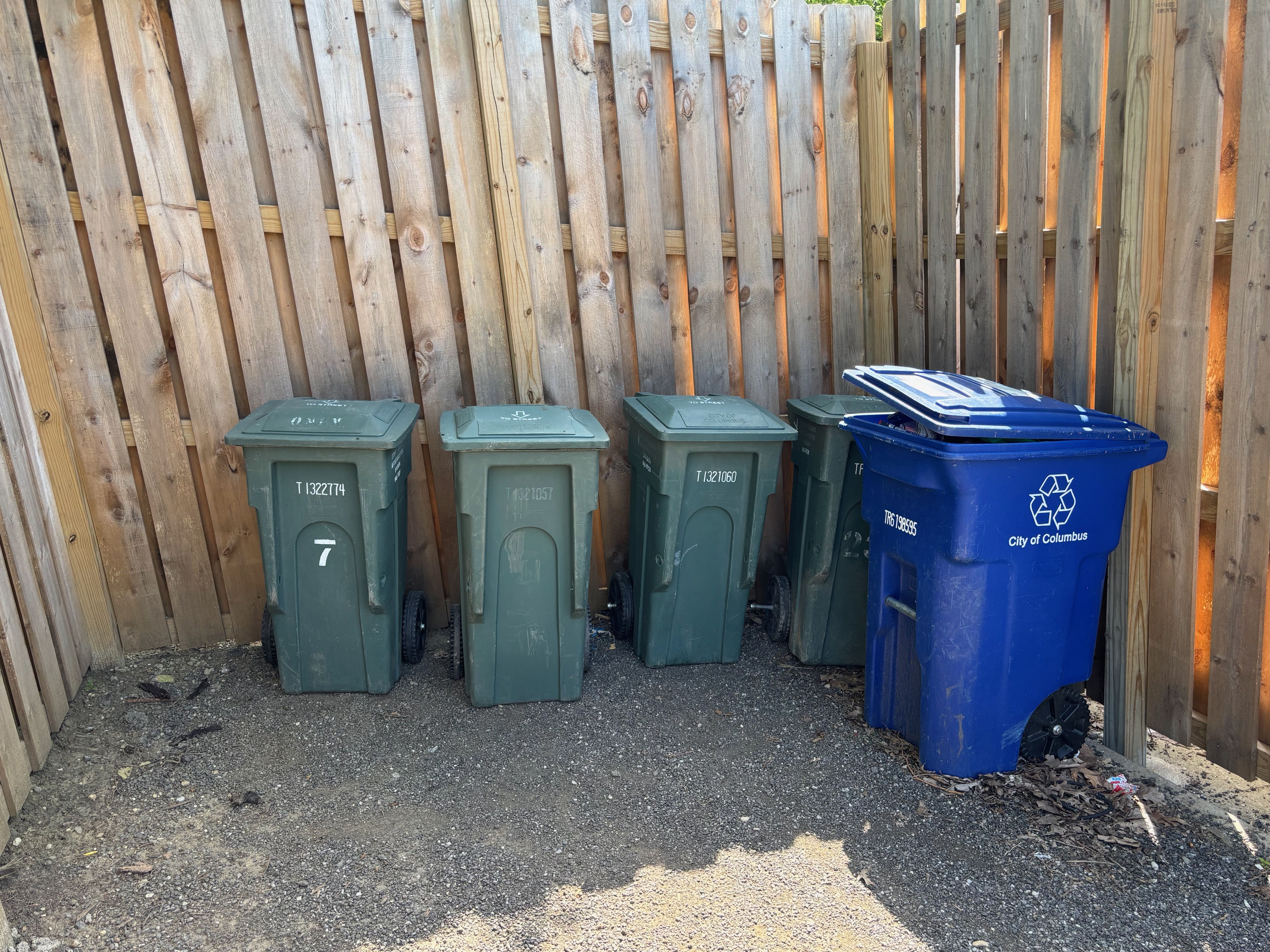 Several wheeled garbage cans (green) and a blue recycling bin sit against a weathered wooden fence on a gravel lot; sunlight glints through the gaps in the slats.