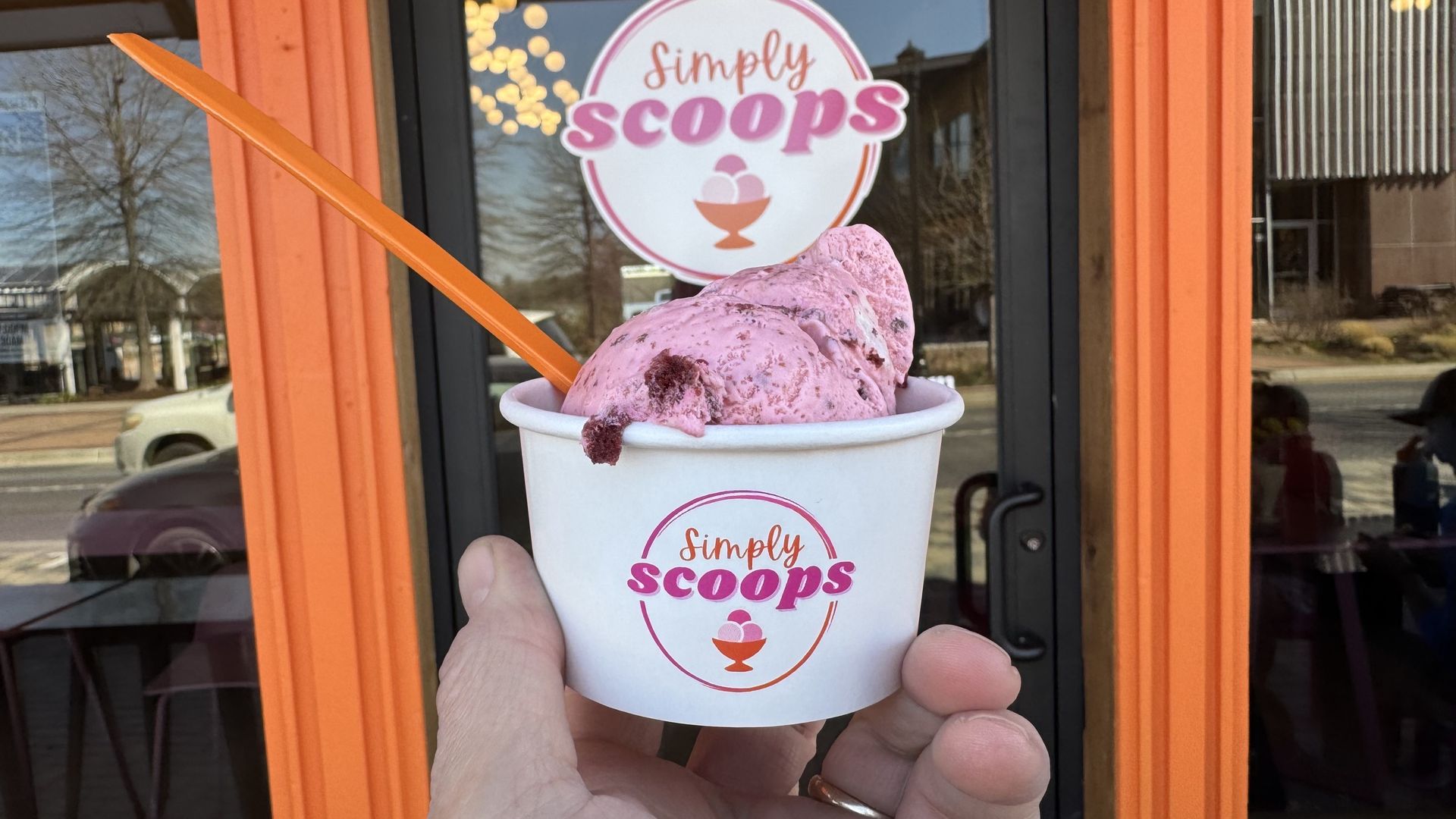 A hand holds a white cup with pink ice cream and an orange spoon, in front of an orange storefront window; a circular sign behind reads "Simply Scoops" in pink letters.