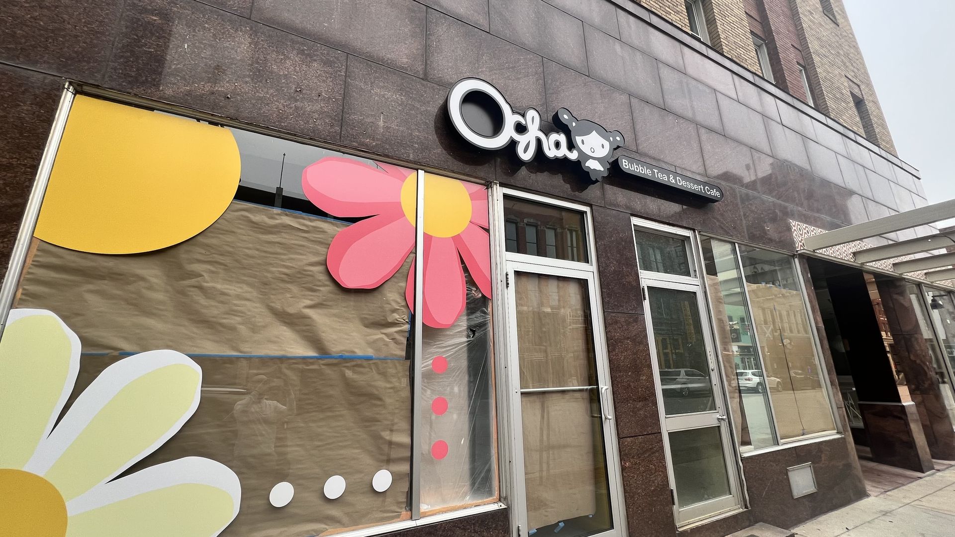 Storefront of Ocha Bubble Tea & Dessert Cafe with boarded windows decorated with oversized yellow and pink flowers and white dots on brown paper.