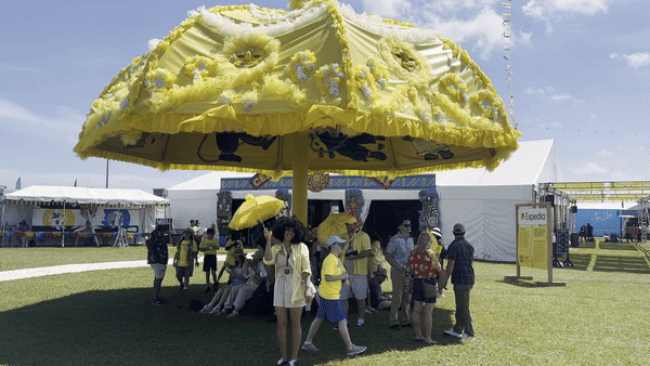 A woman with a small yellow umbrella dances below a large, sculptural yellow umbrella.
