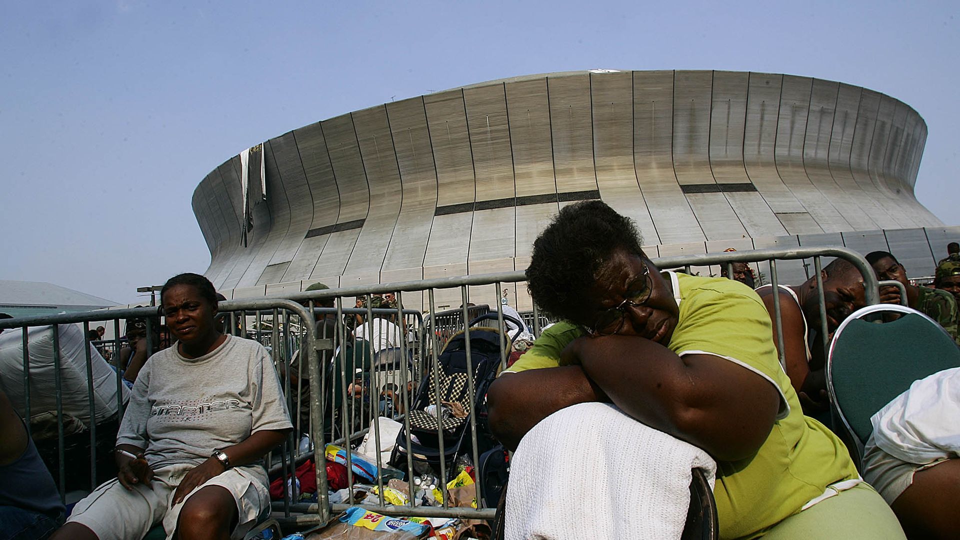 People sitting and resting near metal barricades outside a large, curved stadium under a clear sky, surrounded by bags and supplies, some appearing tired or distressed.